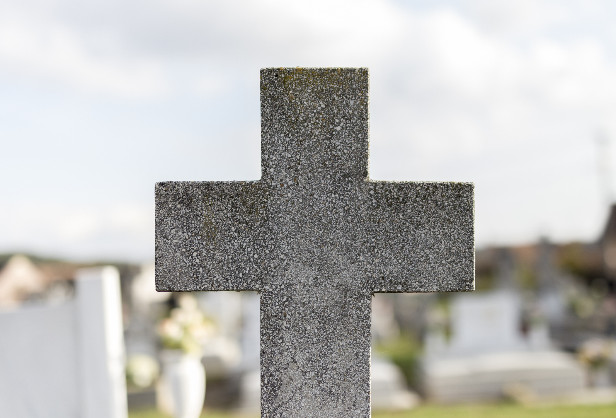 Gravestone cross at a cemetery with several blurry headstones and flowers in the background