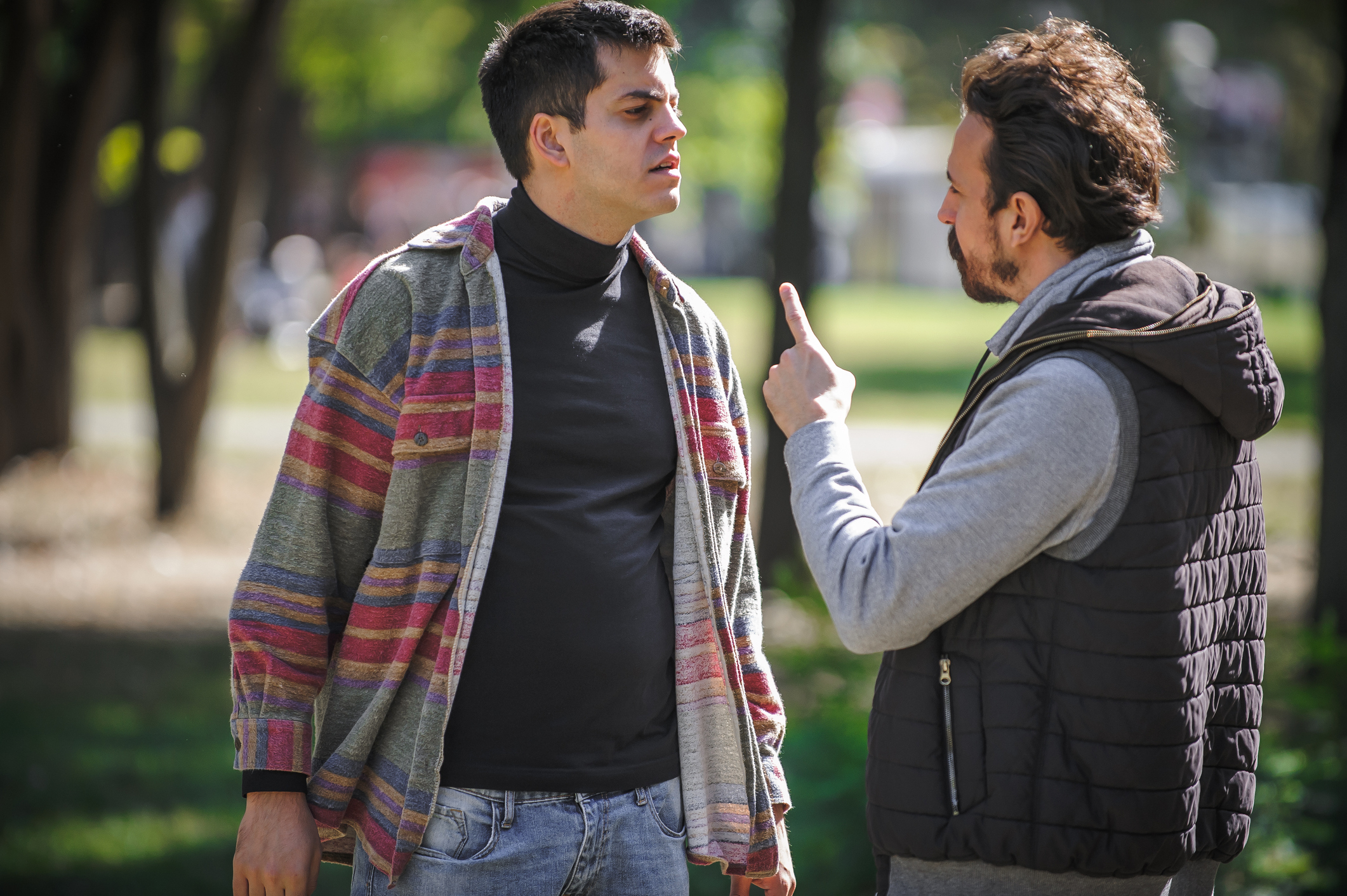 Two men argue in a park. One man wears a striped cardigan and black turtleneck, while the other wears a gray long-sleeve and black vest, pointing a finger