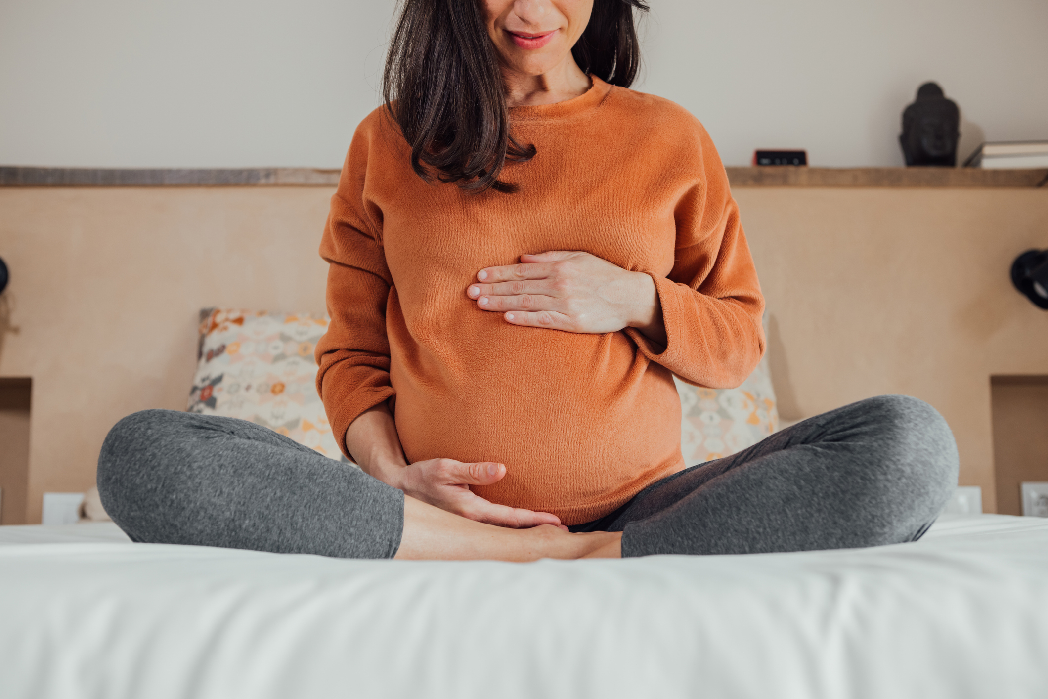 A pregnant woman sits cross-legged on a bed, gently cradling her belly with both hands
