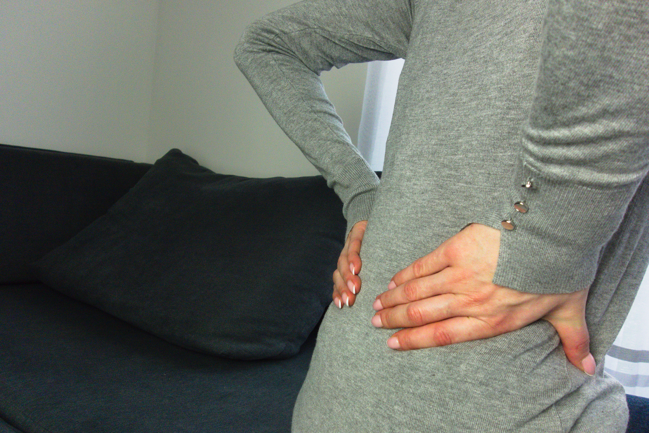 Person in a grey outfit holding their lower back with both hands, likely due to discomfort or pain