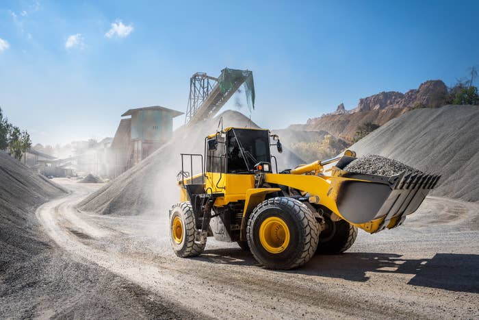 A large yellow excavator transports gravel at an industrial site with piles of gravel and a conveyor belt in the background