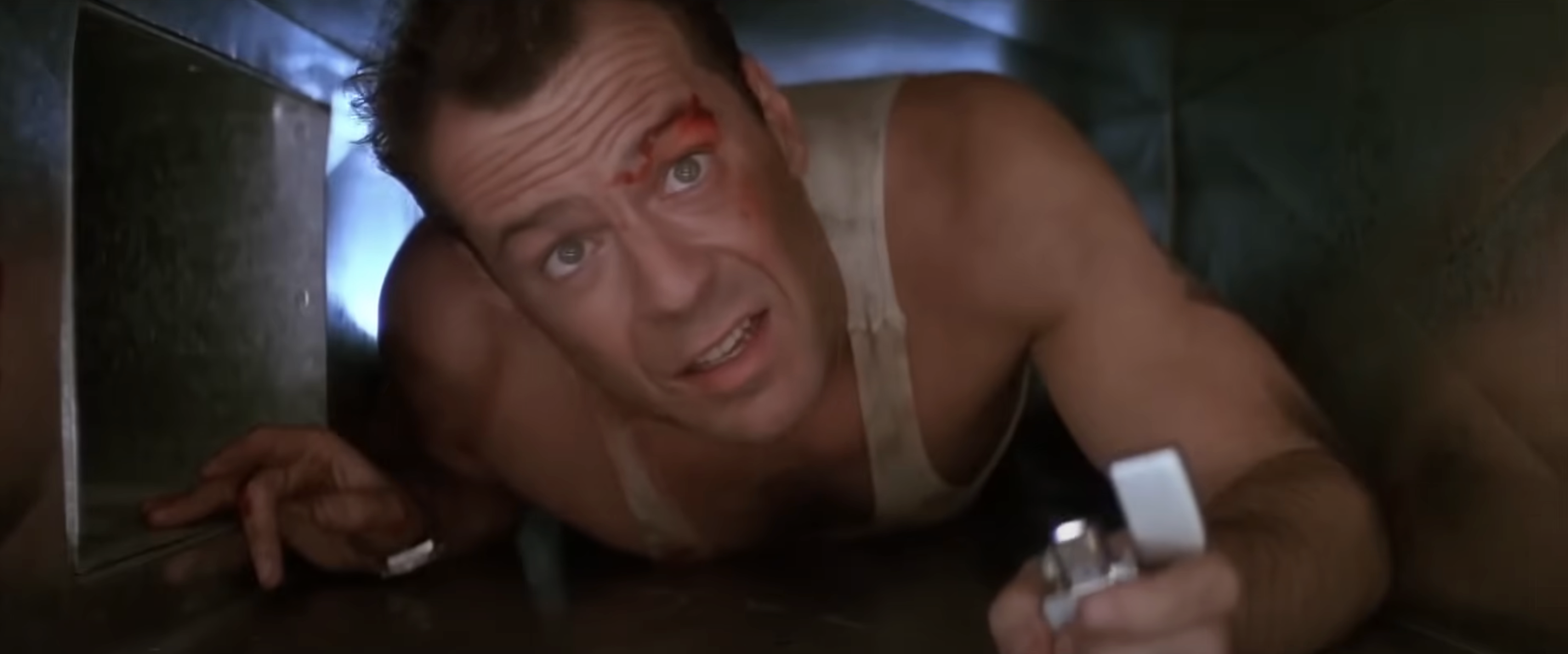 Bruce Willis in a tank top, crawling in a narrow air vent, looking forward with a lighter in his hand. He has a small wound on his forehead
