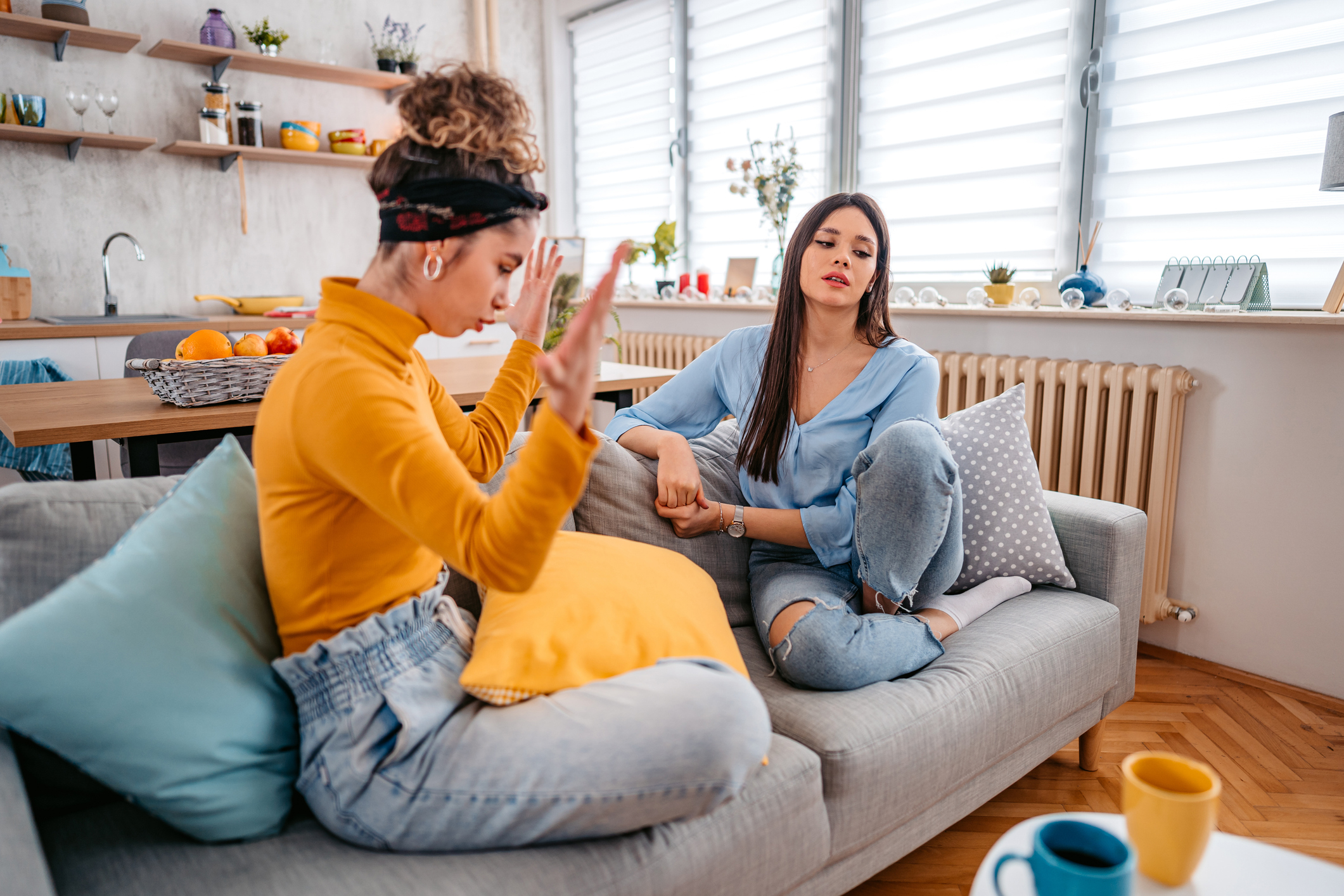 Two women, one raising her hand and the other sitting with a concerned expression, are having a conversation on a couch in a modern living room