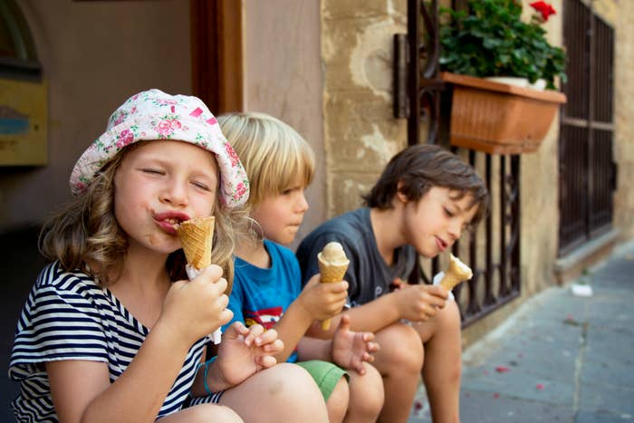 Three children, two boys and a girl wearing a floral hat, sit on a stone step licking ice cream cones, enjoying a warm day outdoors