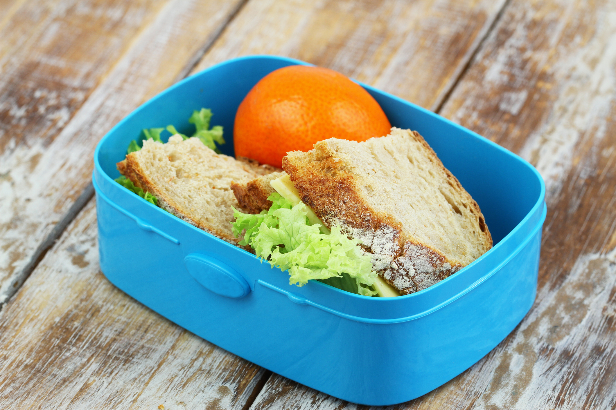 A blue lunchbox with a sandwich made of leafy lettuce and thick bread, alongside a whole orange, placed on a wooden surface
