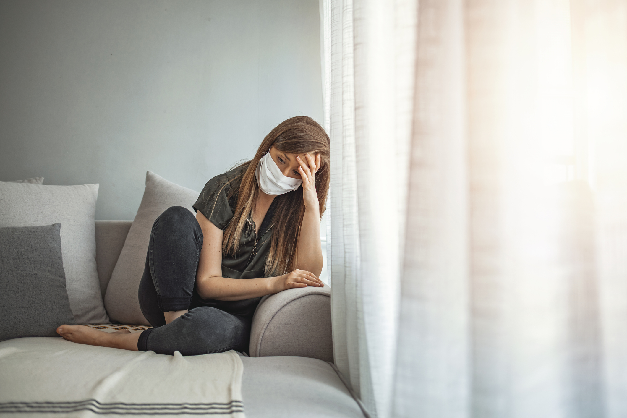 A person wearing a mask sits on a couch, appearing distressed with their head resting on their hand, while looking out of a window with sheer curtains