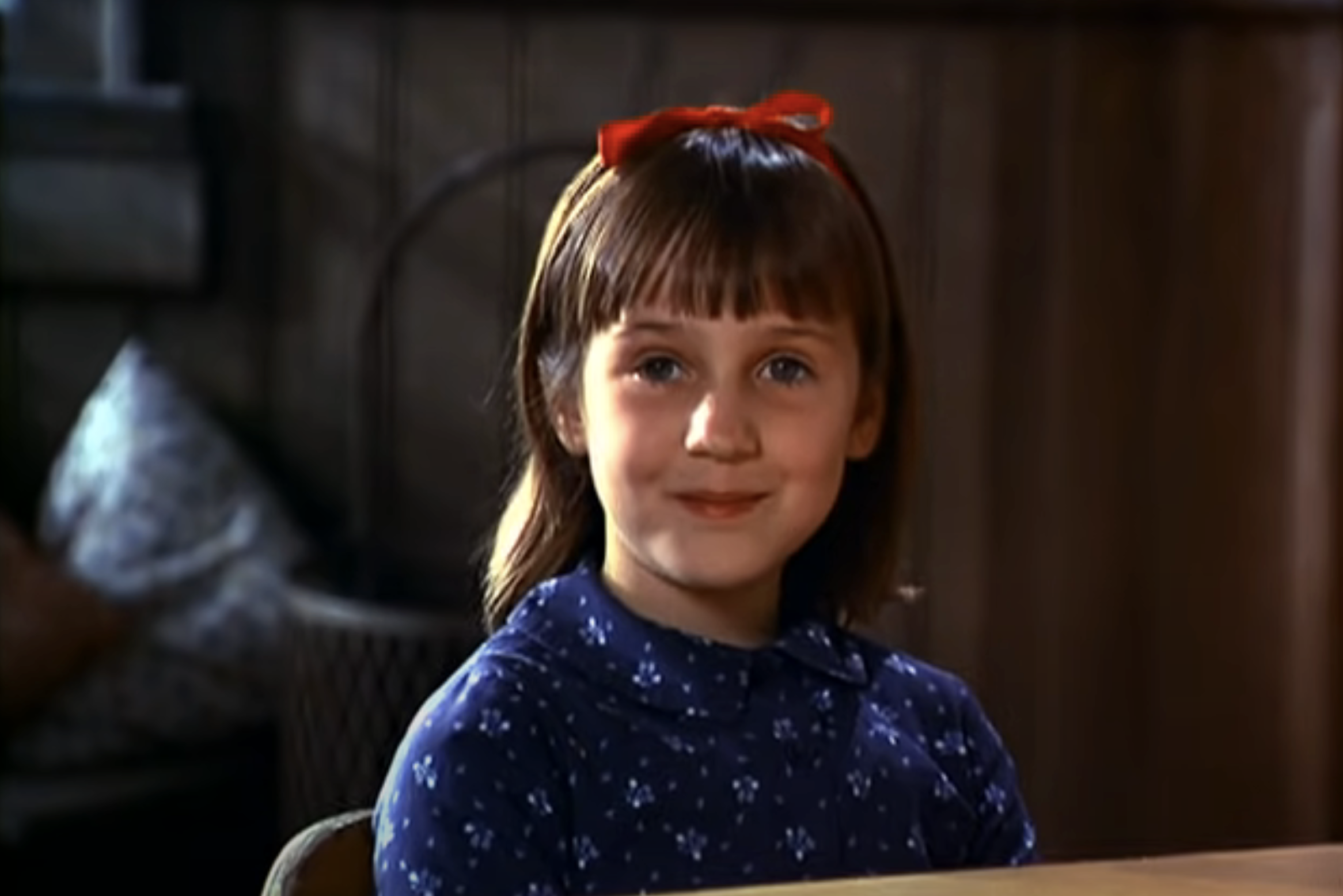 Mara Wilson sits at a table smiling, wearing a polka-dotted dress and a red bow in her hair