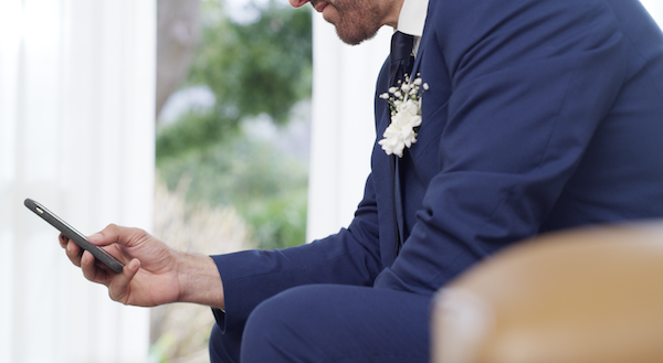 A man in a suit looks at his phone while seated. A small floral boutonniere is pinned to his jacket