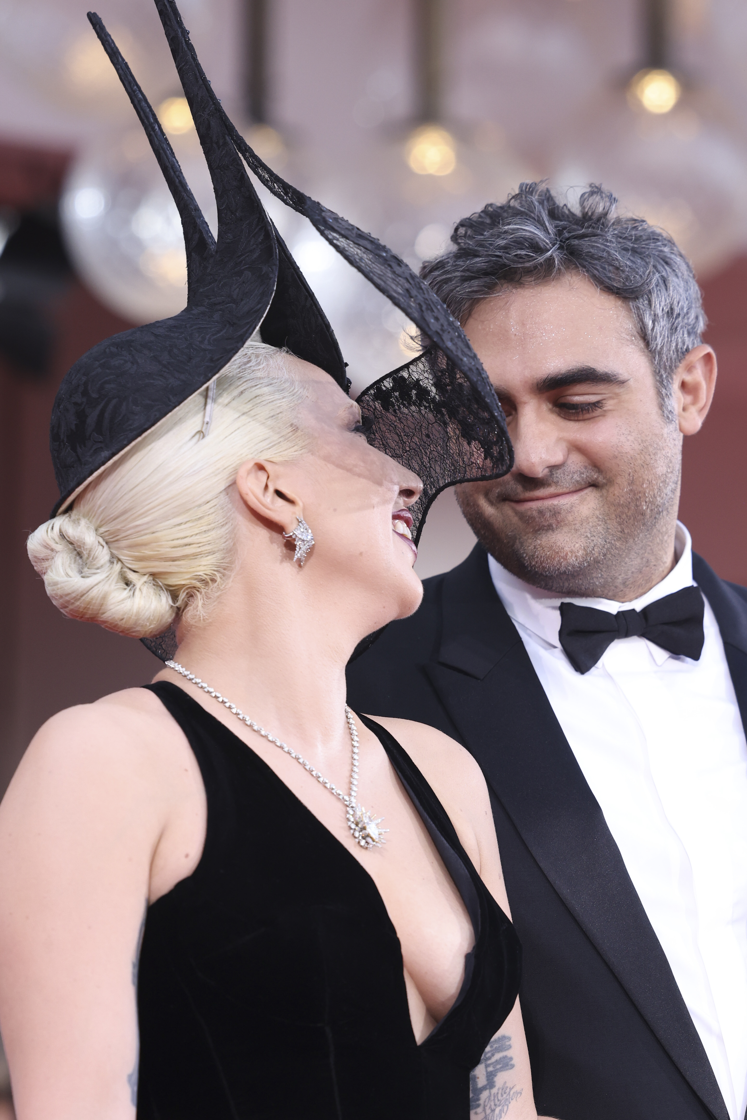 Lady Gaga wears a dramatic headpiece and elegant black dress, stands close to a man in a black tuxedo, both smiling at each other on the red carpet