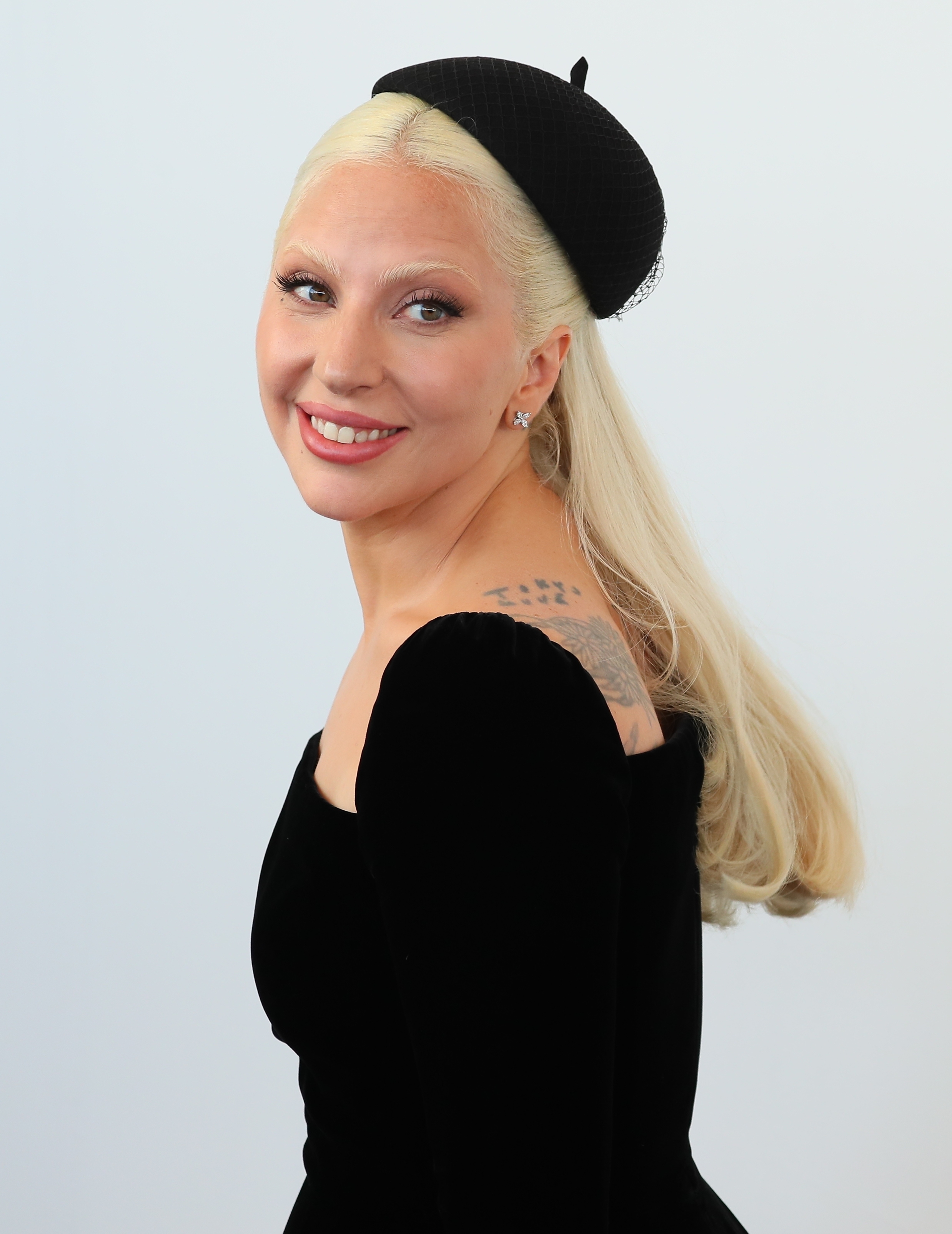 Lady Gaga wears a black dress with a matching headpiece, smiling and showing a shoulder tattoo