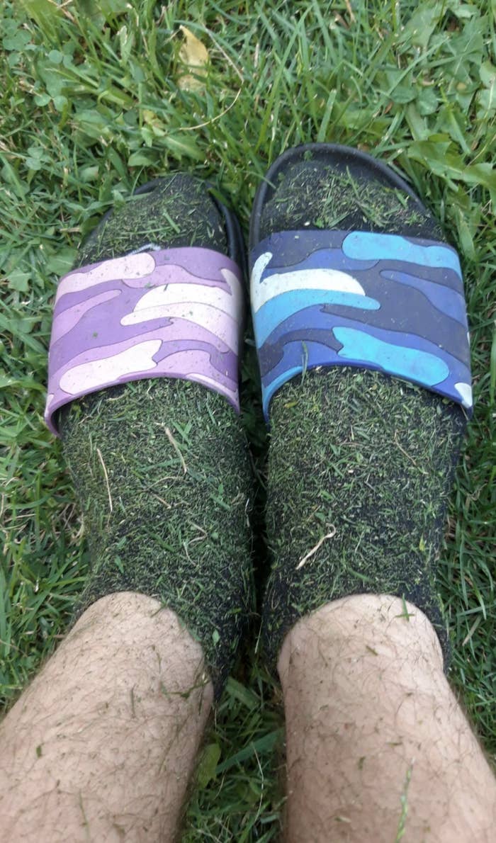 Two feet in grass, wearing mismatched sandals (one purple camouflage, one blue camouflage) and socks covered in grass clippings