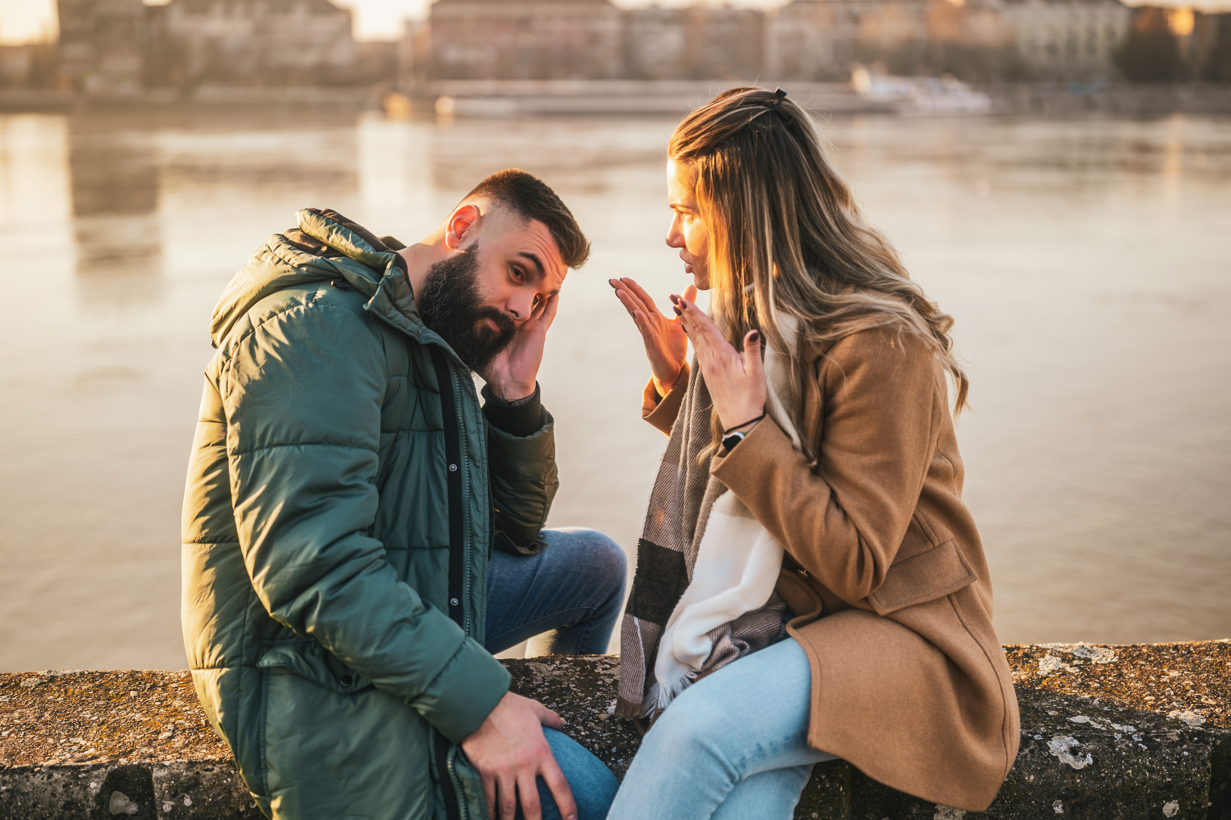 A man and woman are engaged in a heated conversation while sitting by the water. The woman appears frustrated, gesturing with her hands, while the man looks upset