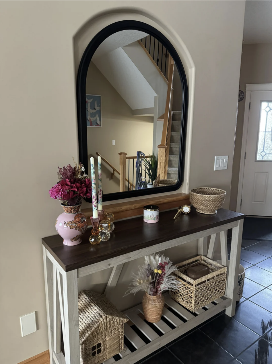 A home entryway features a decorative console table with floral arrangements, candles, woven baskets, and a mirror that's too small for the section on the wall