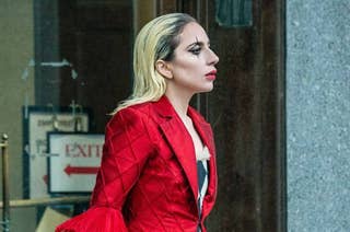 Lady Gaga wearing a stylish, red, quilted jacket with pleated sleeves, walking outside a building. An EXIT sign is partially visible in the background