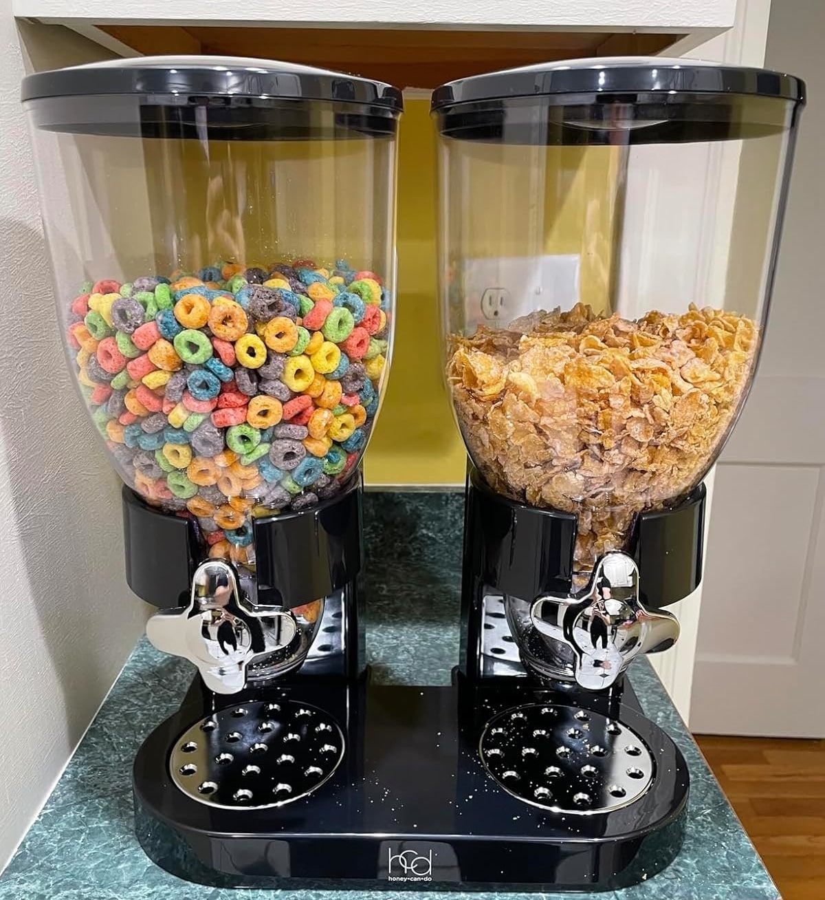 Dual cereal dispensers on a kitchen counter, one filled with colorful loop cereal and the other with cornflakes. Dispensers have silver levers