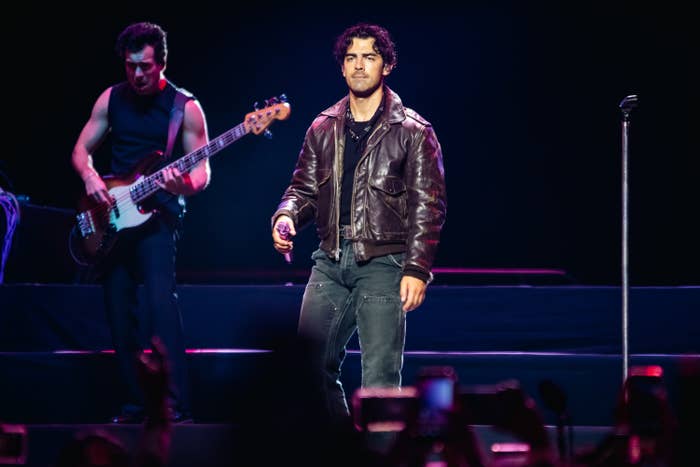 Joe Jonas on stage holding a microphone, wearing a leather jacket and dark pants, with a bassist playing in the background