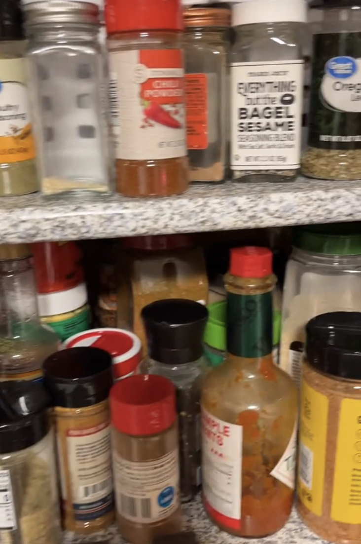 Various spice jars and seasoning containers arranged on two shelves in a pantry, including brands like Everything But The Bagel Sesame, Cholula, and Old Bay