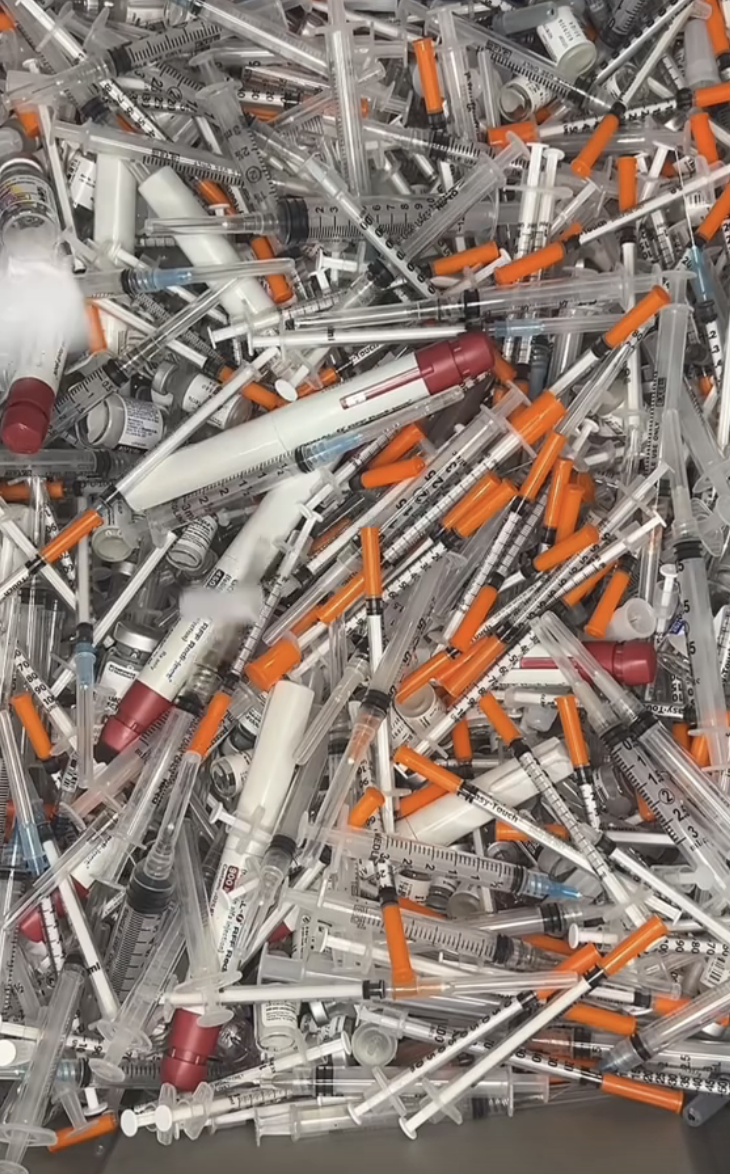 A large pile of various used medical syringes and injection vials, appearing as medical waste