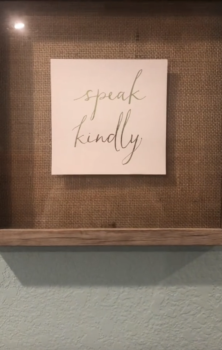 A framed sign on a wall reads, "speak kindly." The sign is placed against a burlap background with a wooden shelf below it