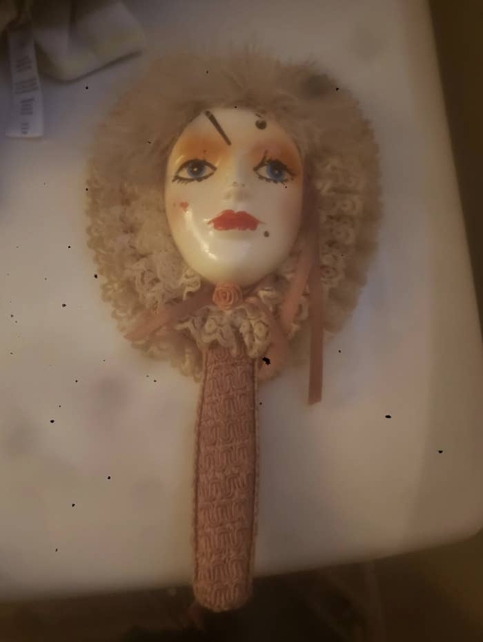 A hand mirror with a doll face on the back, featuring painted facial details, a crochet bonnet, and ribbon accents