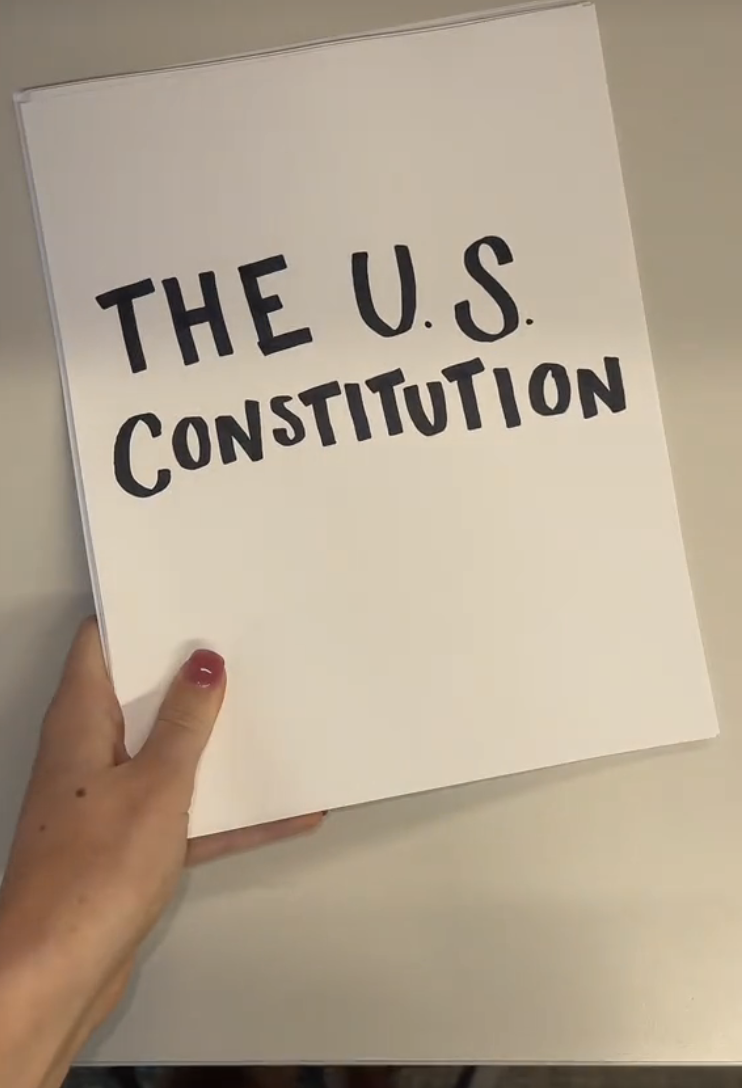 A hand holding a document titled "The U.S. Constitution" in bold letters