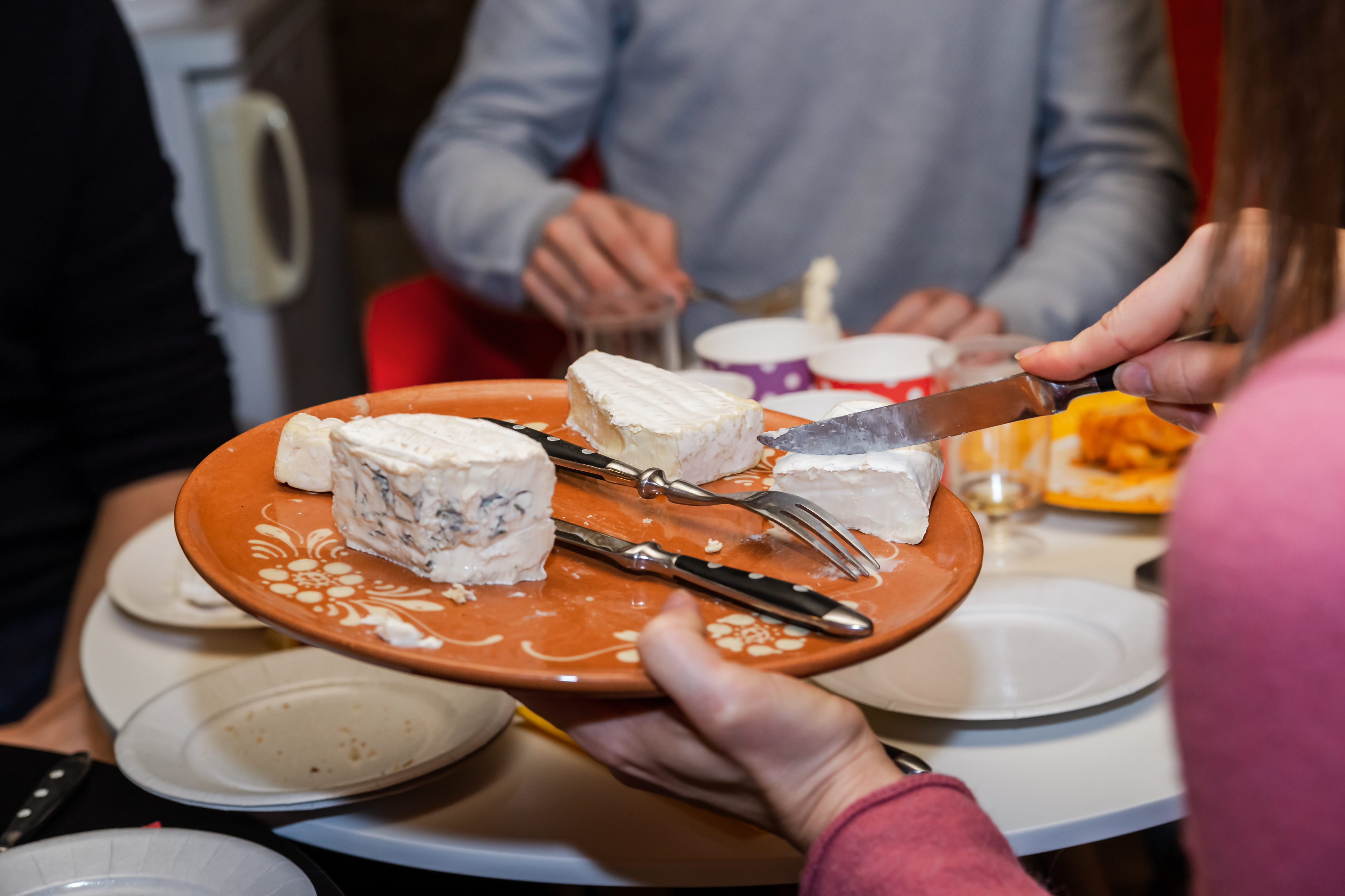 A person serves a plate of assorted cheeses at a dining table with three people, whose identities are unknown, seen sitting and reaching for the food