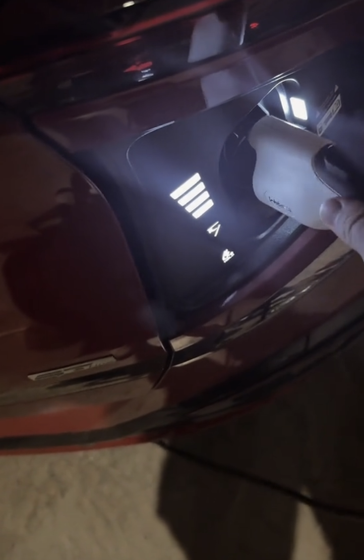 A person's hand plugs a charging cable into an electric car at a charging port, which is illuminated in the dark
