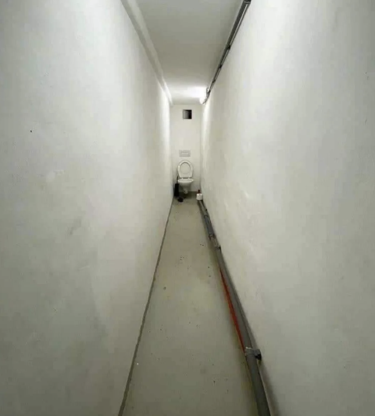 A narrow, empty corridor with a single chair and trash bin at the far end against the back wall. Pipes run along the right side of the corridor