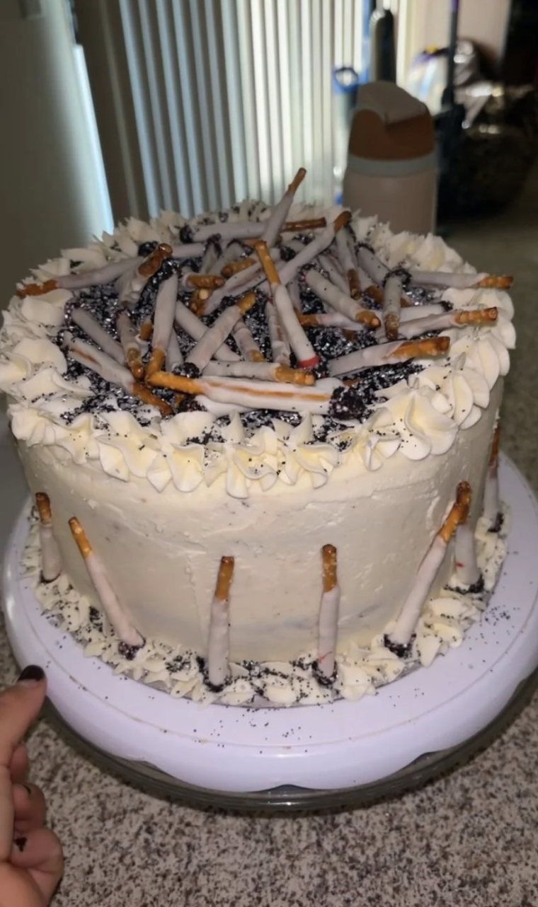A decorated cake with white frosting, topped with pretzels, chocolate dust, and white icing resembling lit cigarettes. Pretzels are also around the sides