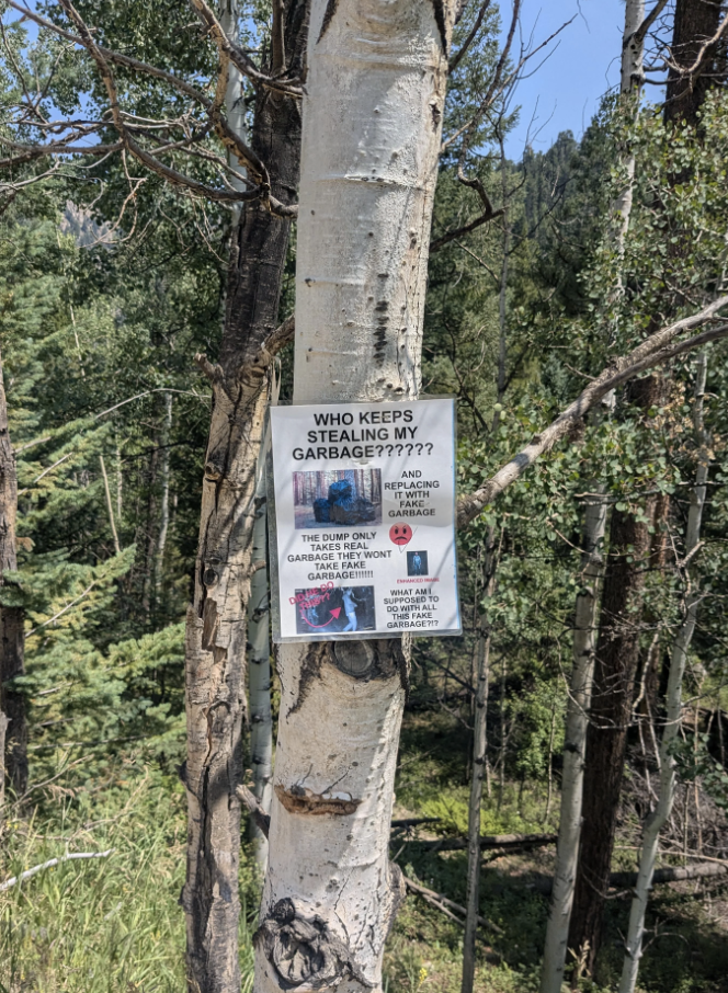 Sign taped to a tree in a forest asking, "Who keeps stealing my garbage????" and stating that the dump only accepts garbage, not recyclables