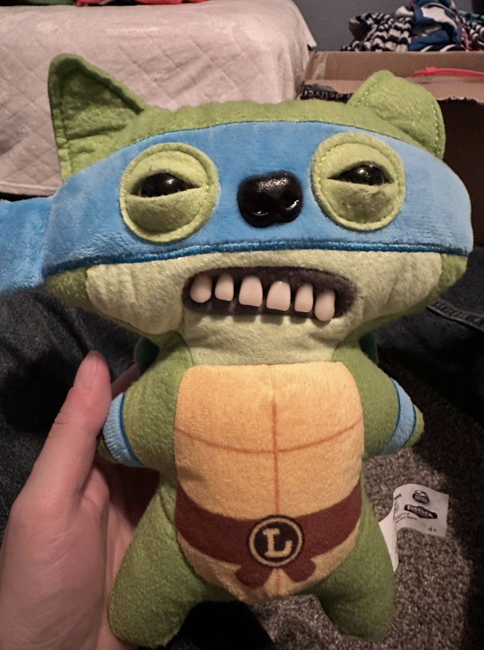 Person holding a plush toy resembling a cartoon character with a blue mask, large eyes, and big teeth