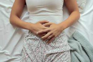 A woman's hands are resting on her abdomen as she lies on a white bed. She is wearing a white tank top and patterned pants