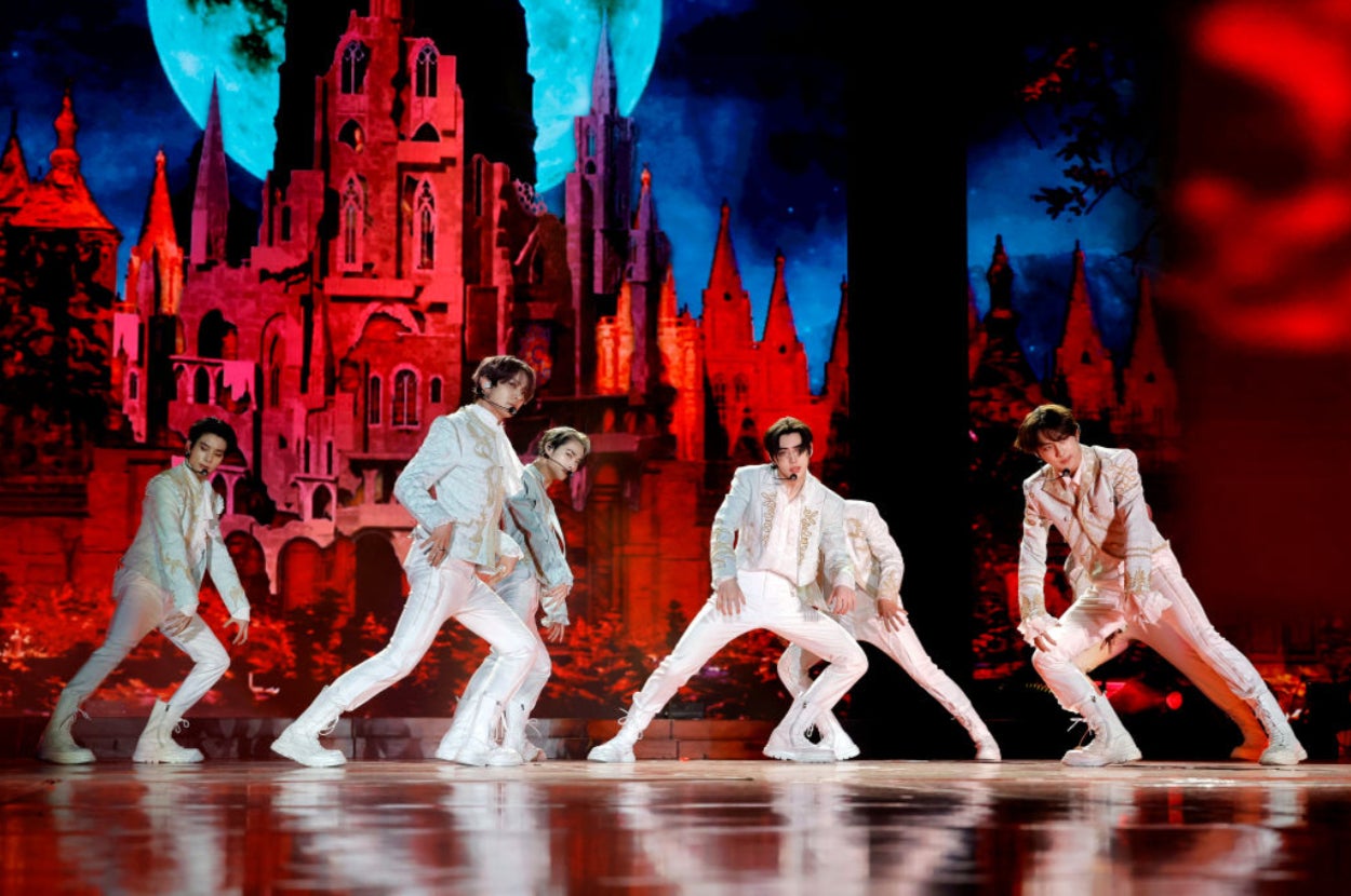 Six performers dance energetically on stage in white outfits, with a dramatic castle backdrop. The environment is theatrical.
