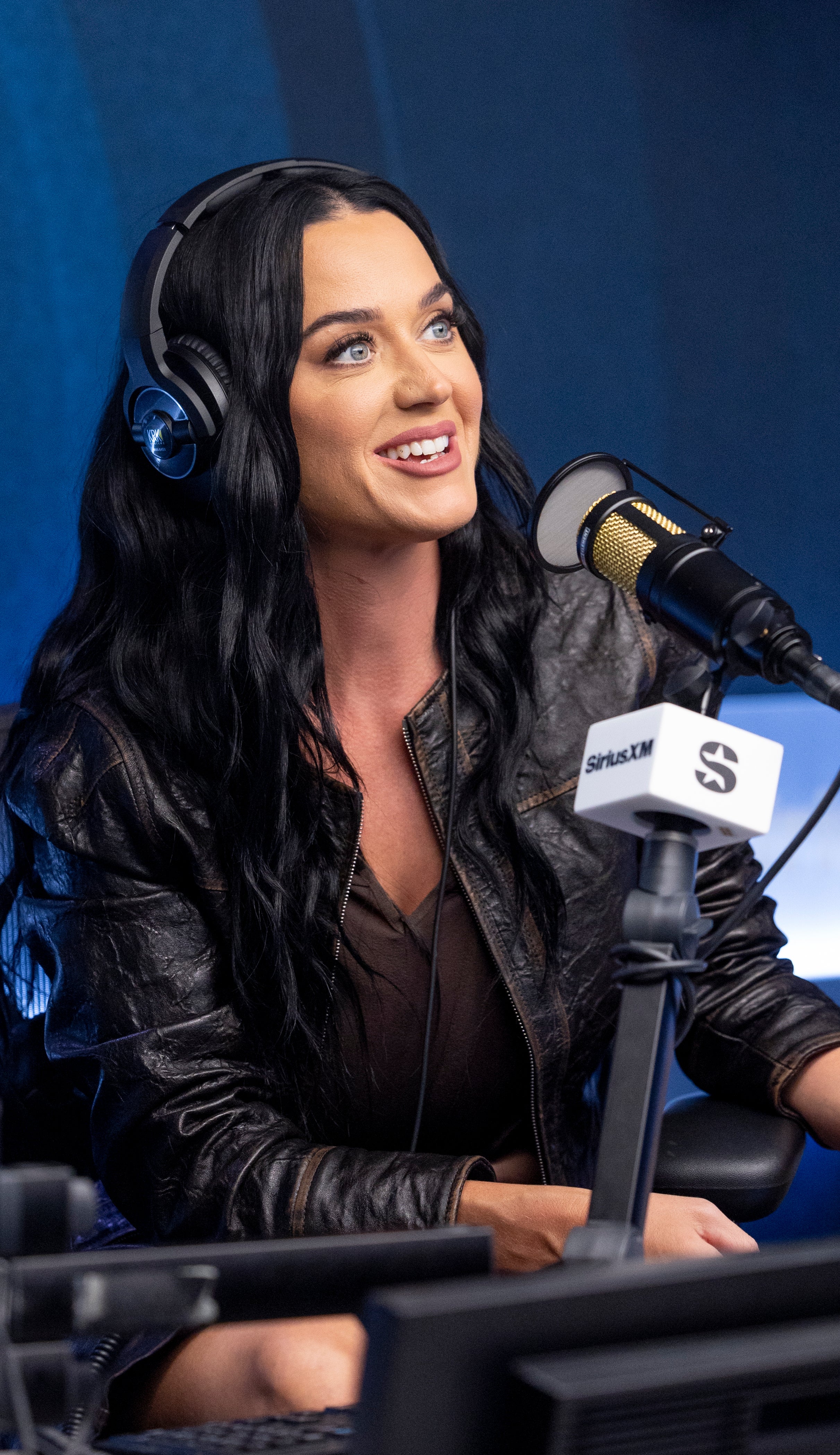 Katy Perry in a radio studio, smiling and speaking into a microphone, wearing headphones and a leather jacket