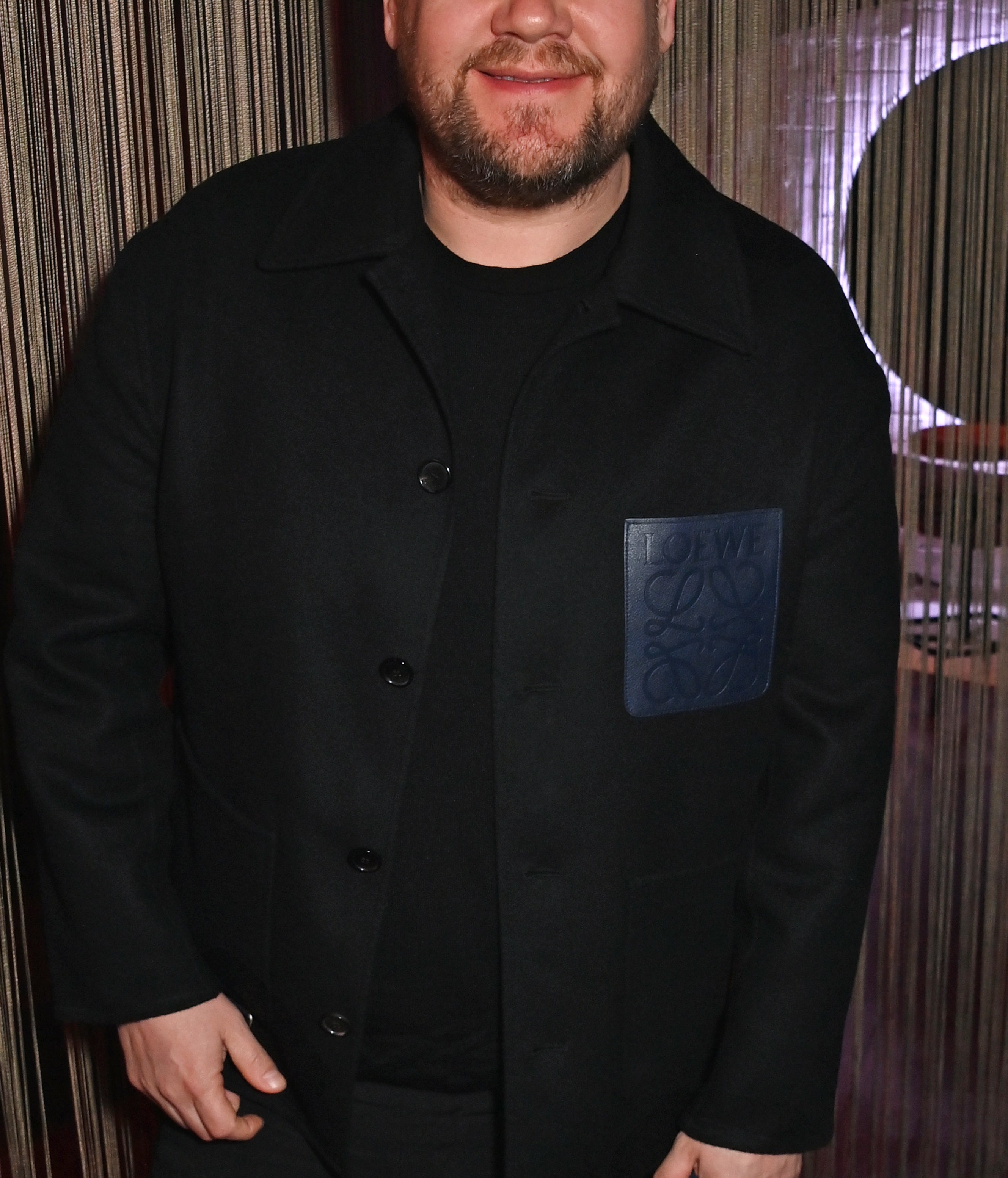 James Corden smiling while posing in a dark outfit with one hand in a pocket and the other holding an object, standing against a string curtain backdrop