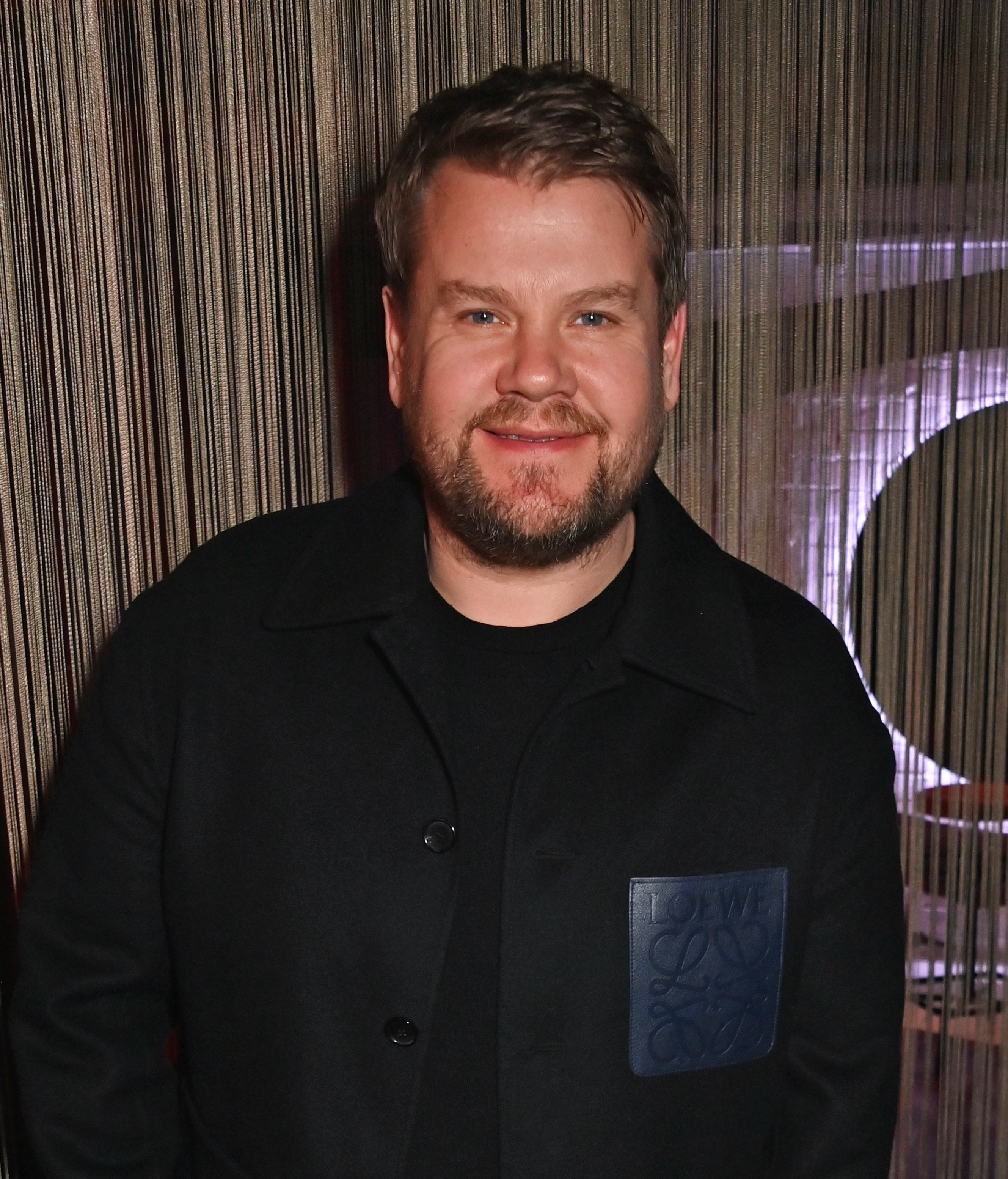 James Corden smiling while posing in a dark outfit with one hand in a pocket and the other holding an object, standing against a string curtain backdrop