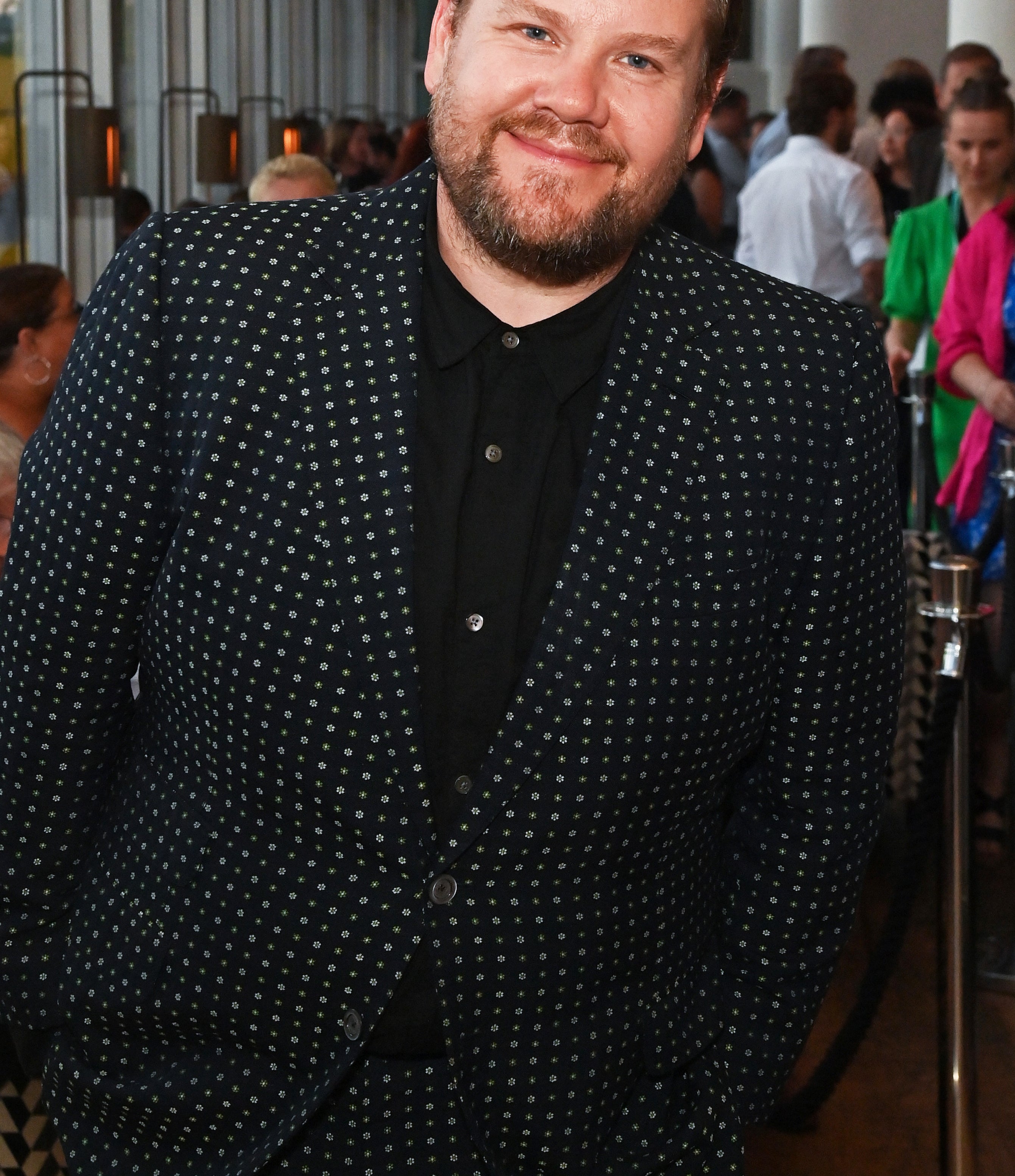 James Corden is smiling, wearing a polka-dot suit and shirt at an event with attendees in the background