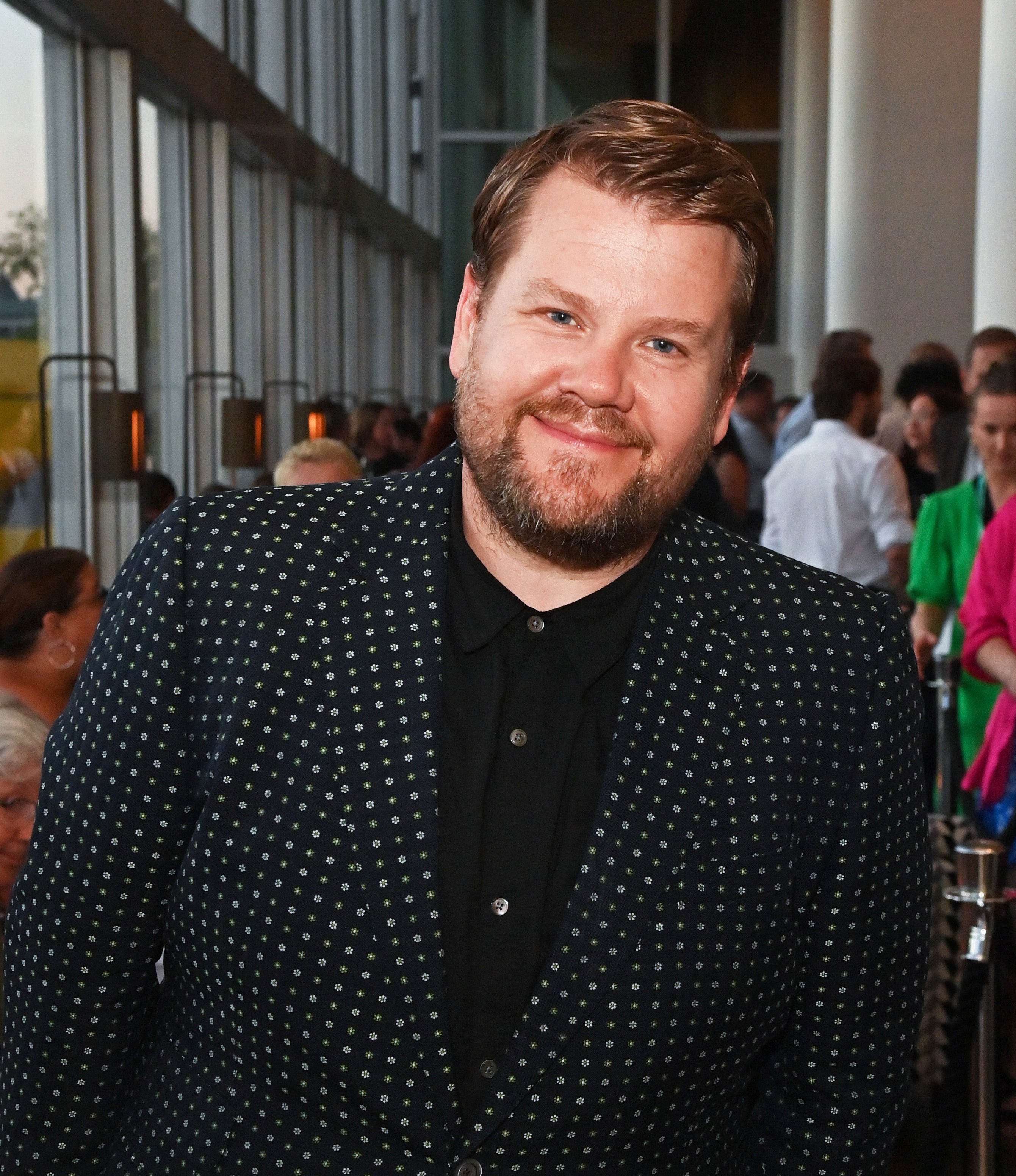James Corden is smiling, wearing a polka-dot suit and shirt at an event with attendees in the background
