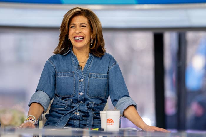 Hoda Kotb smiles while seated at a desk, wearing a denim shirt and hoop earrings, with a "Today" show mug in front of her