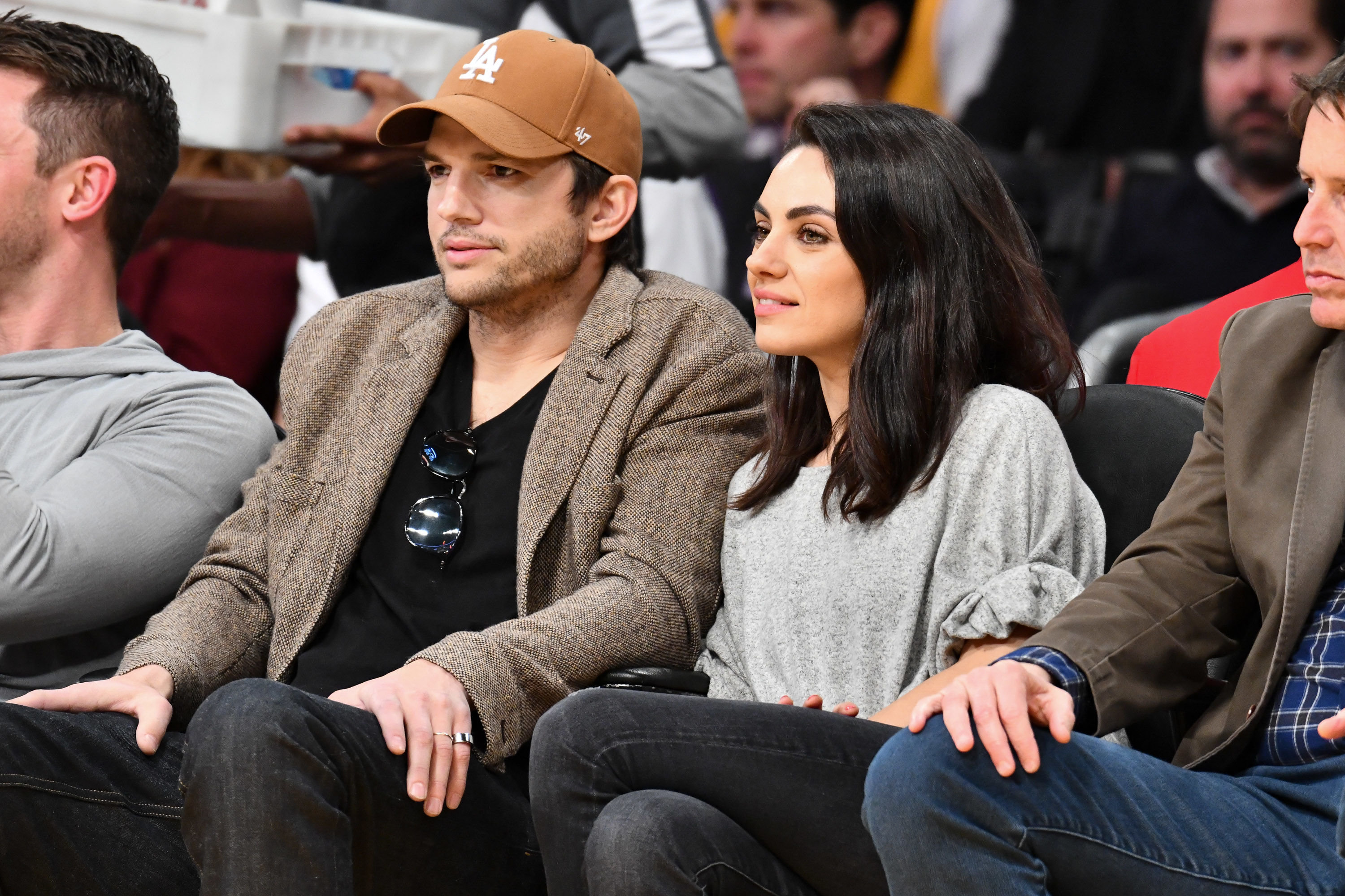 Ashton Kutcher in a blazer and baseball cap and Mila Kunis in a casual top seated at an event