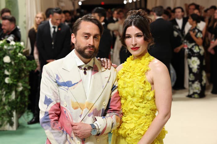 Kieran Culkin in a pastel-patterned suit and Jazz Charton in a yellow floral dress on a red carpet, with people in the background
