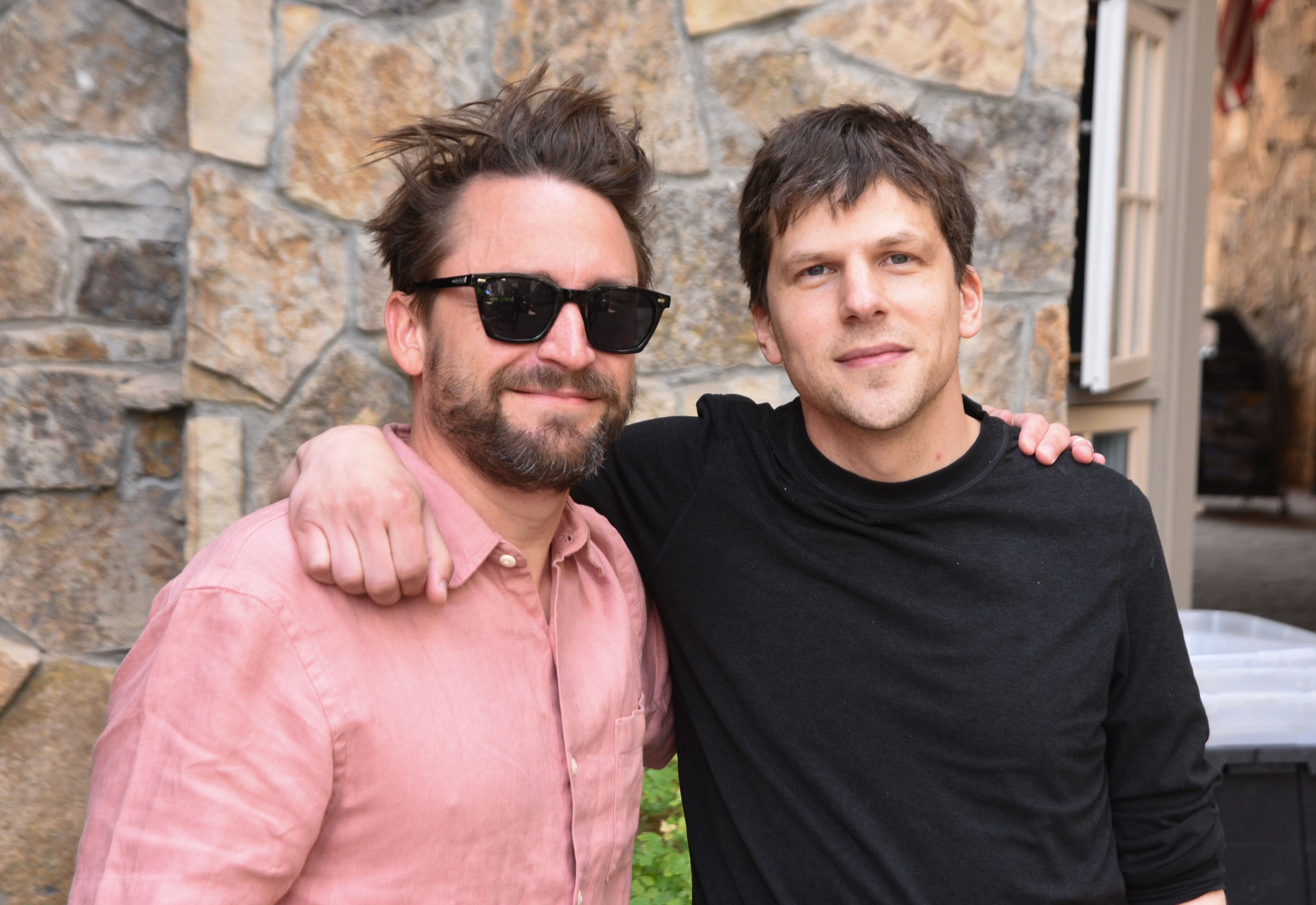 Kieran Culkin and Jesse Eisenberg, dressed in casual shirts, pose together in front of a stone wall. Charlie wears sunglasses and has his arm around Jesse