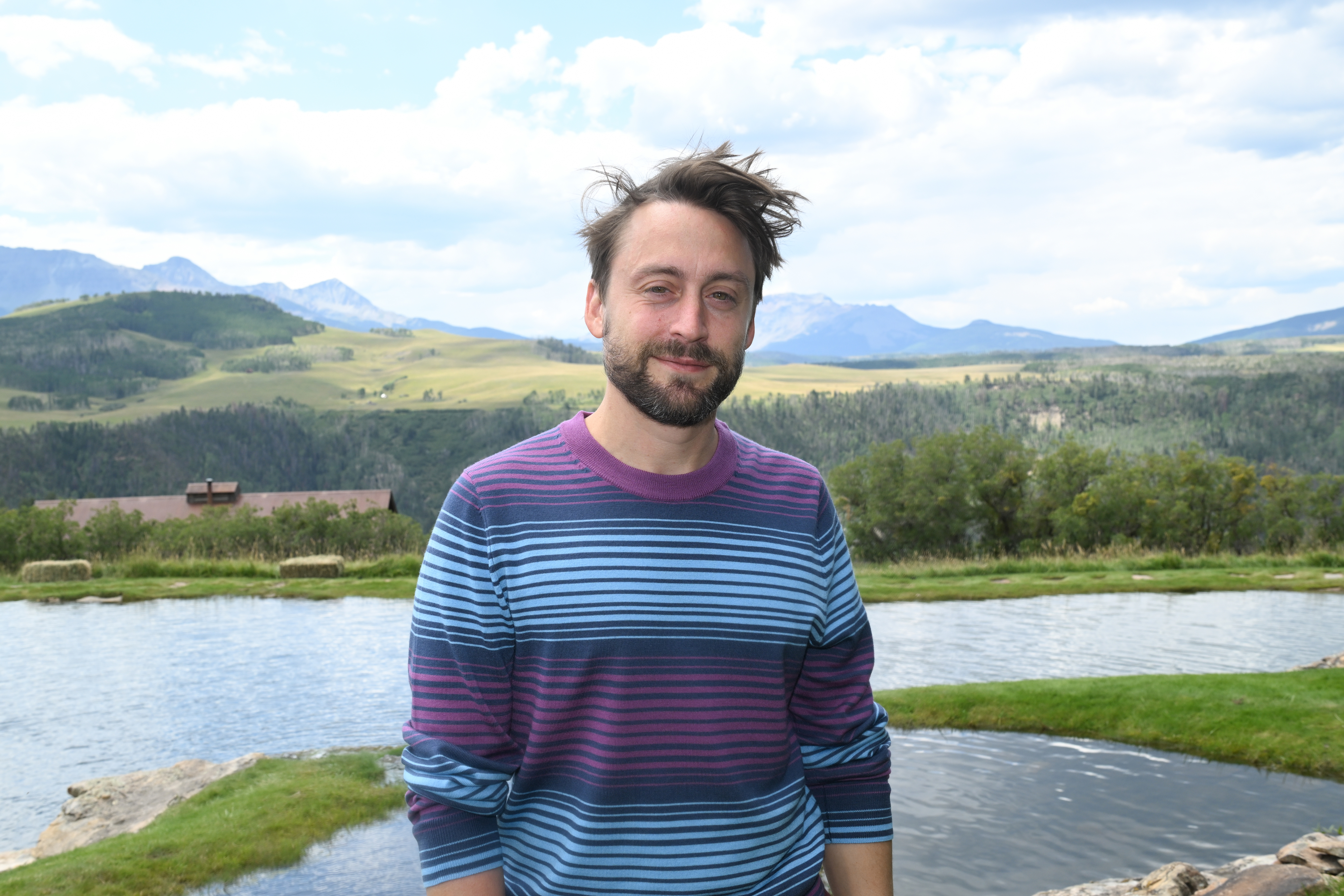 Kieran Culkin stands outdoors in front of a scenic landscape with hills and a pond, wearing a striped long-sleeve shirt