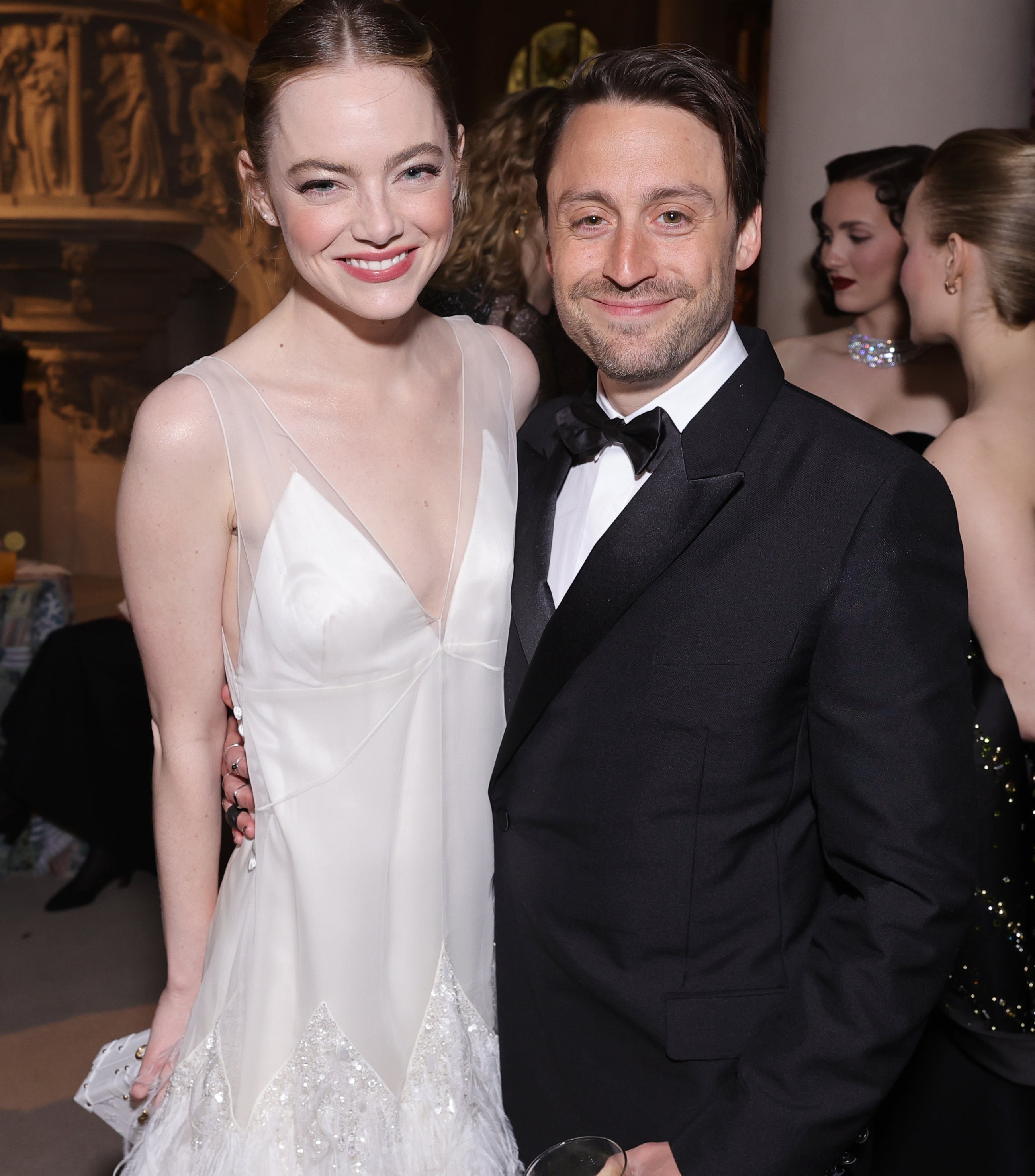 Emma Stone and Kieran Culkin pose together at an event, both smiling; she wears a stylish white dress with feather details, and he wears a black tuxedo