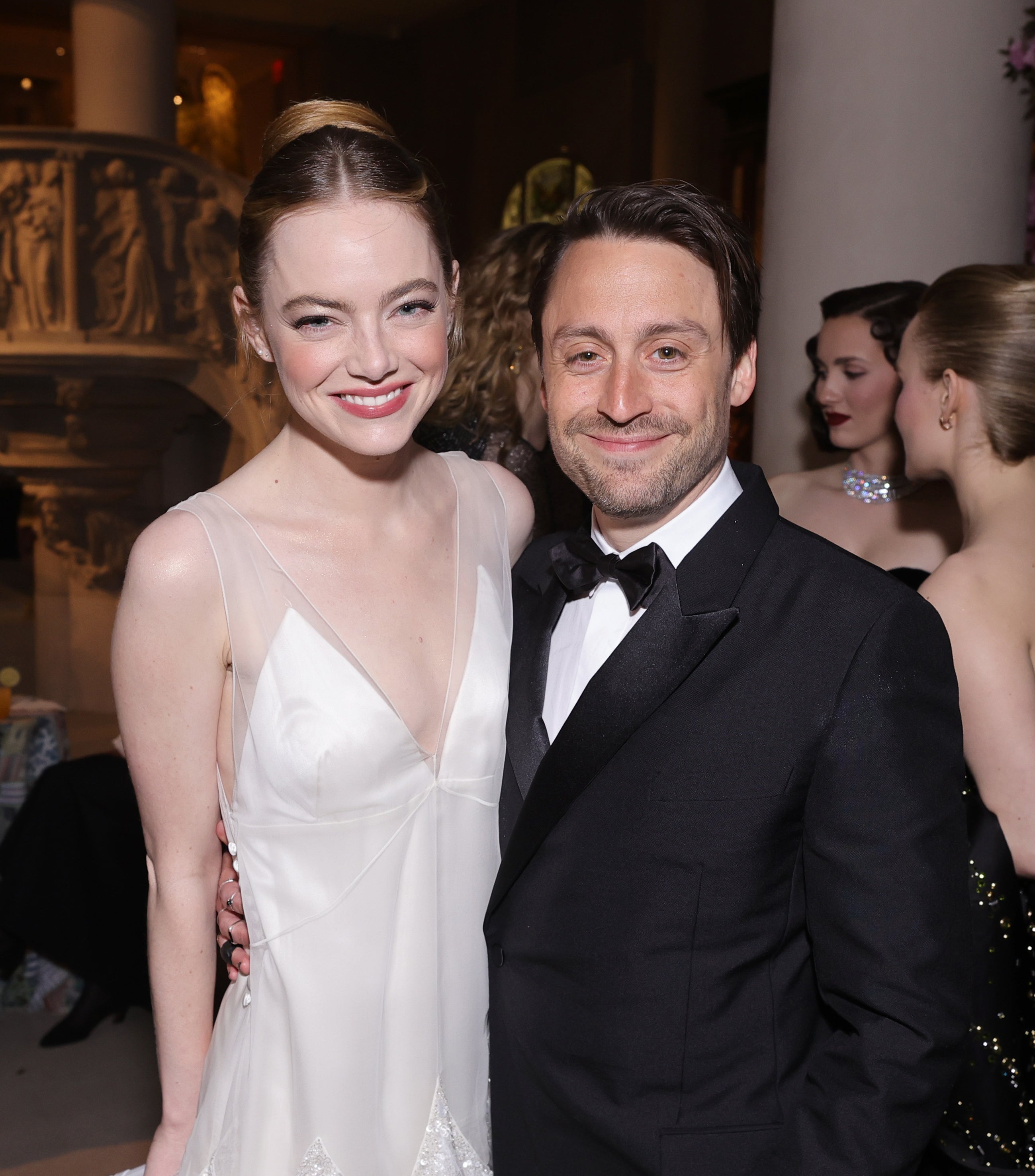 Emma Stone and Kieran Culkin pose together at an event, both smiling; she wears a stylish white dress with feather details, and he wears a black tuxedo