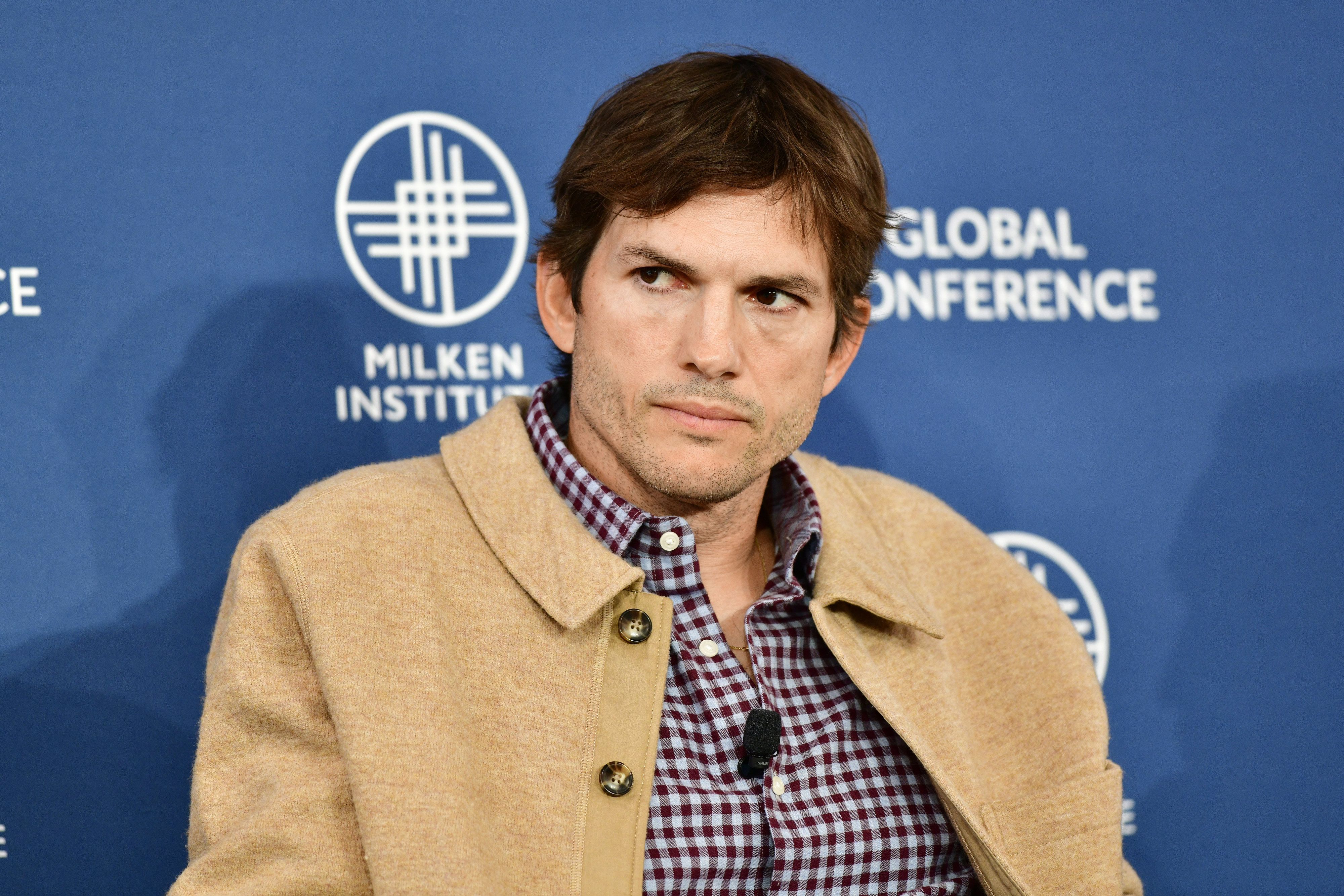 Ashton Kutcher at the Milken Institute Global Conference, wearing a checked shirt and beige jacket, seated in front of a blue backdrop