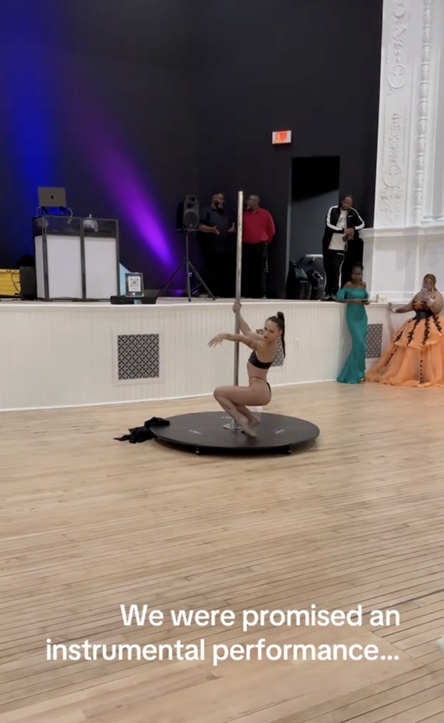 A woman performs a pole dance on a round stage. Spectators, wearing formal attire, stand and watch. The screen text reads, "We were promised an instrumental performance&hellip;"