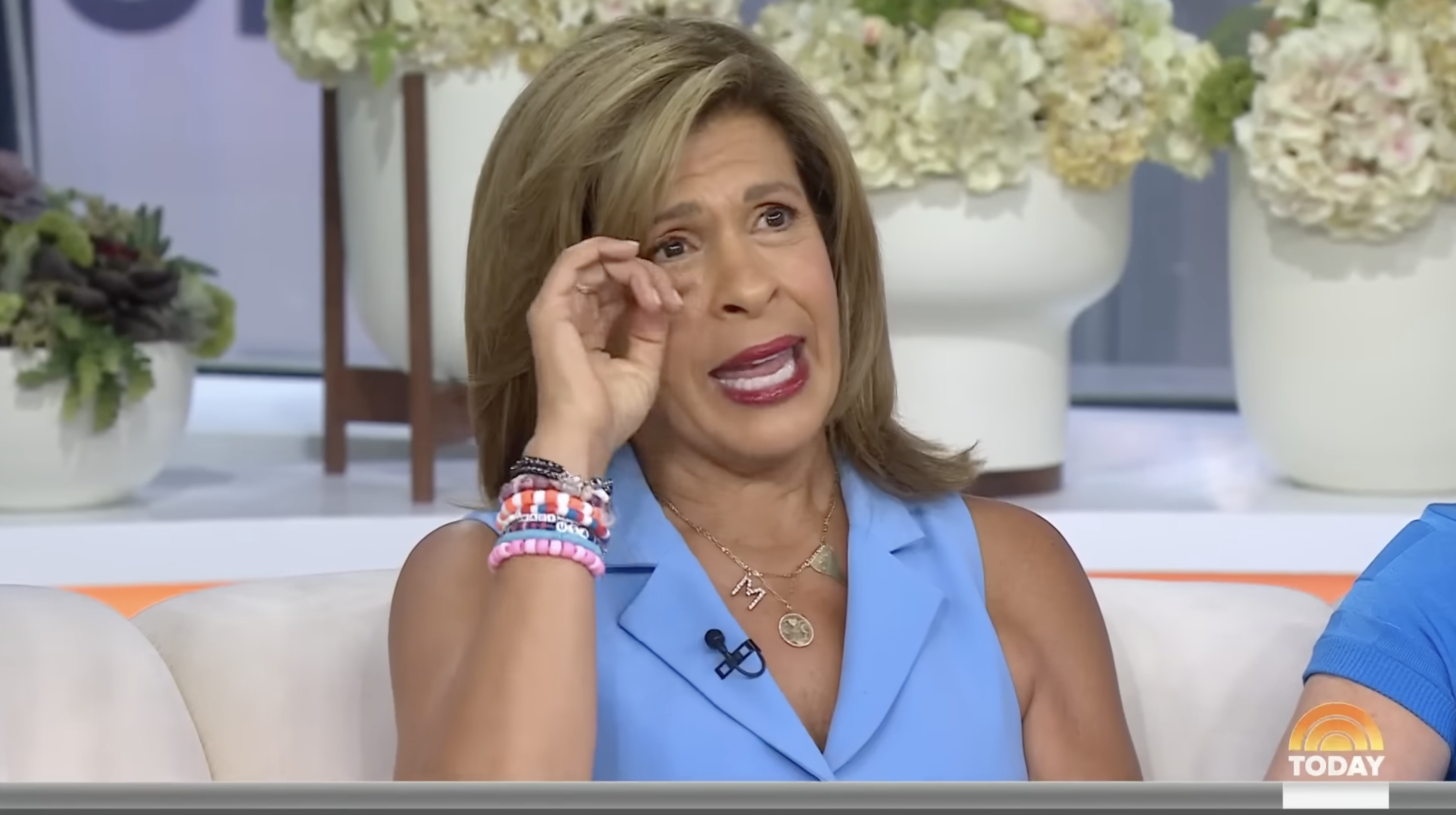 Hoda Kotb on "The Today Show," wearing a sleeveless top with layered bracelets, seated in front of floral arrangements and speaking with tears in her eyes