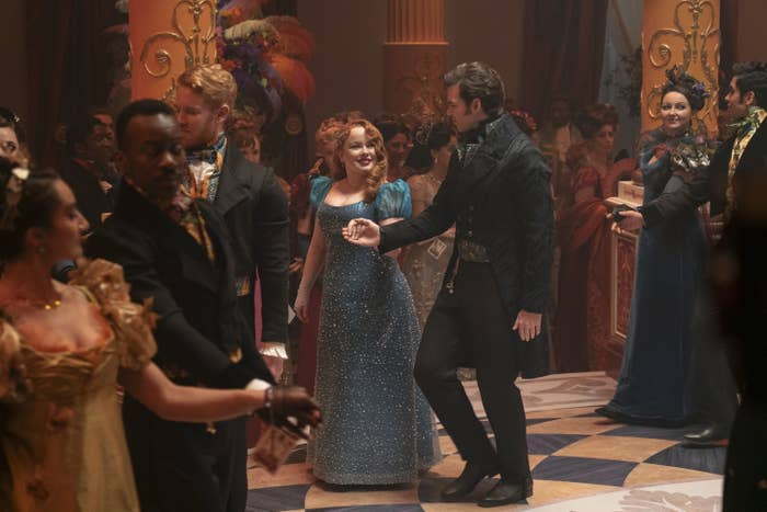Nicola Coughlan and Luke Newton dance together in elegant period costumes at a lavish ballroom event, surrounded by other costumed attendees in a scene from Bridgerton