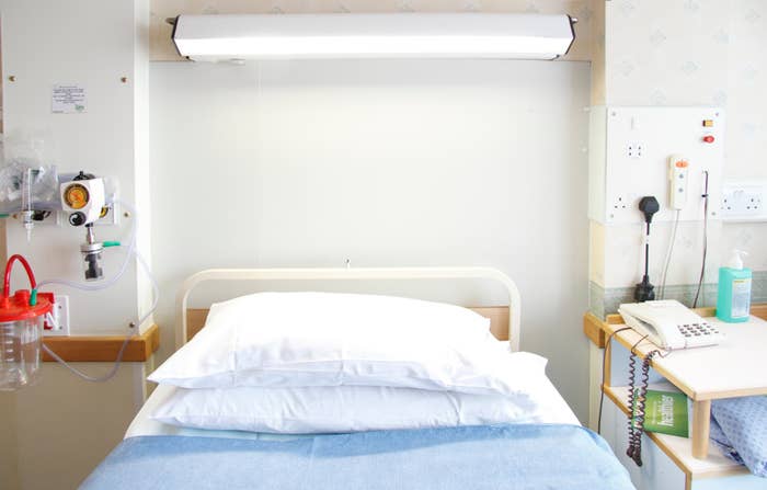 Hospital bed with pillows, an oxygen outlet on the wall to the left, and medical equipment on a side table to the right next to a phone